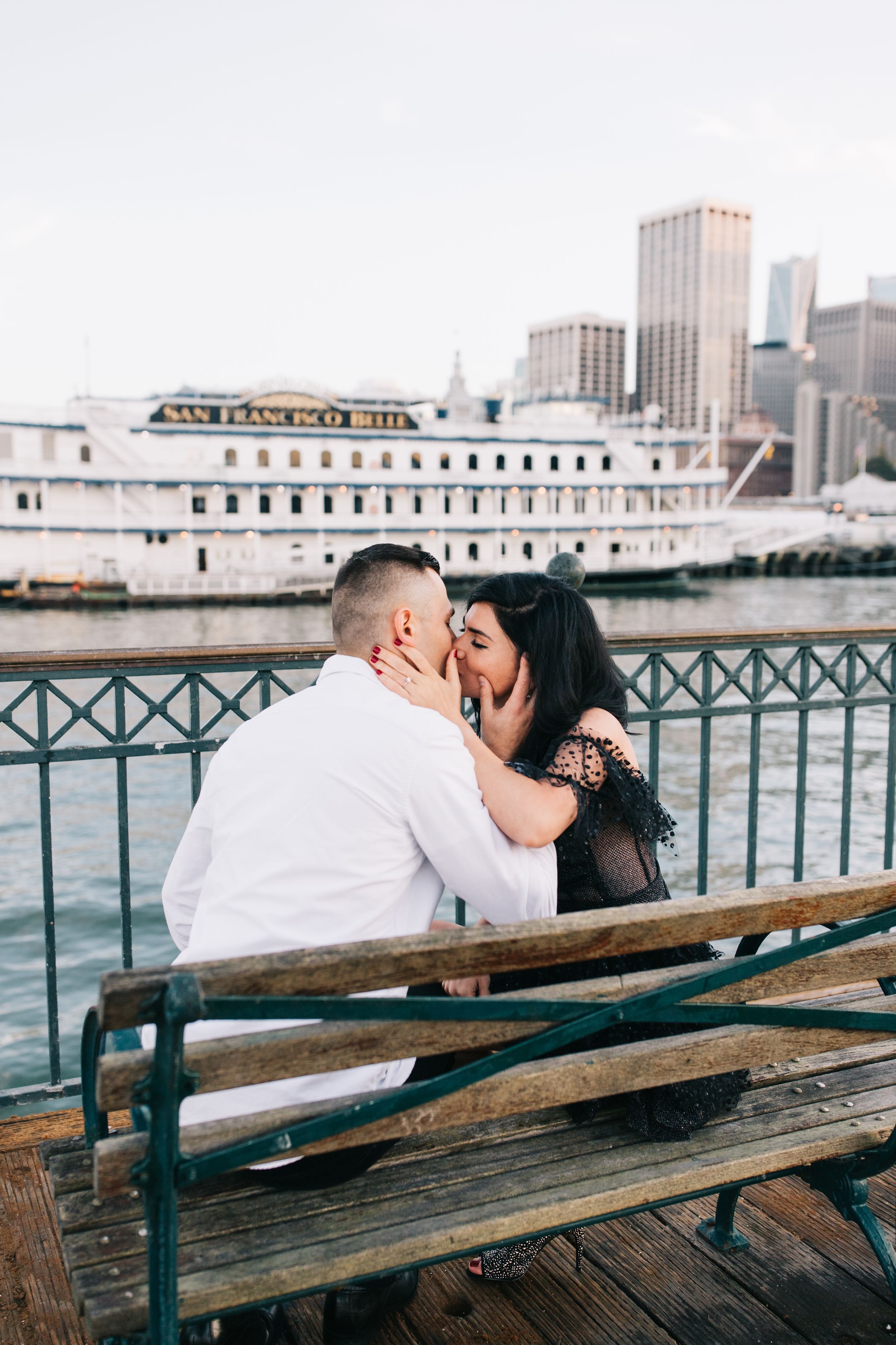 A couple shares a tender moment on a pier bench with a docked cruise ship visible behind them.