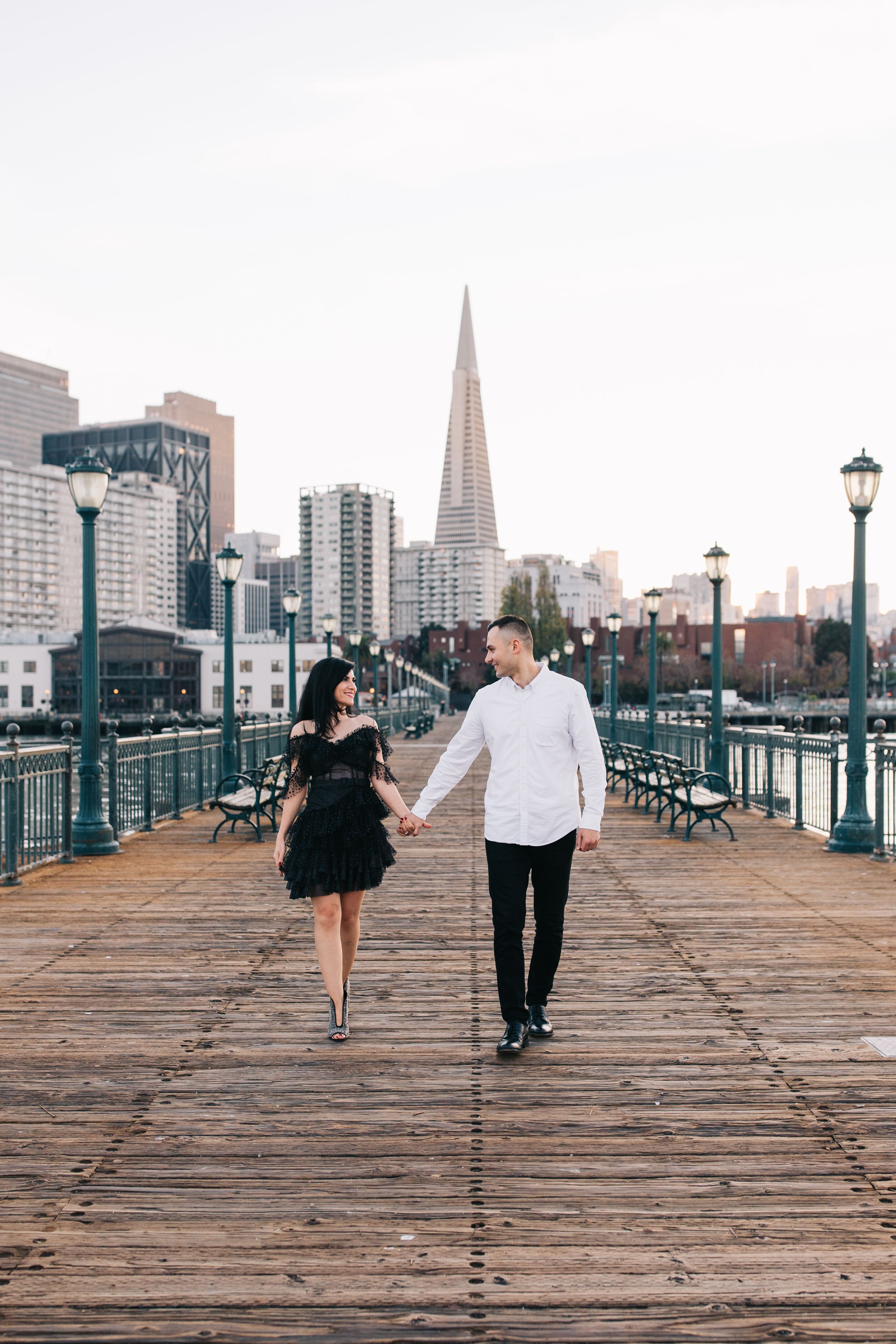 A couple strolls hand in hand down a wooden pier with the iconic San Francisco skyline behind them.