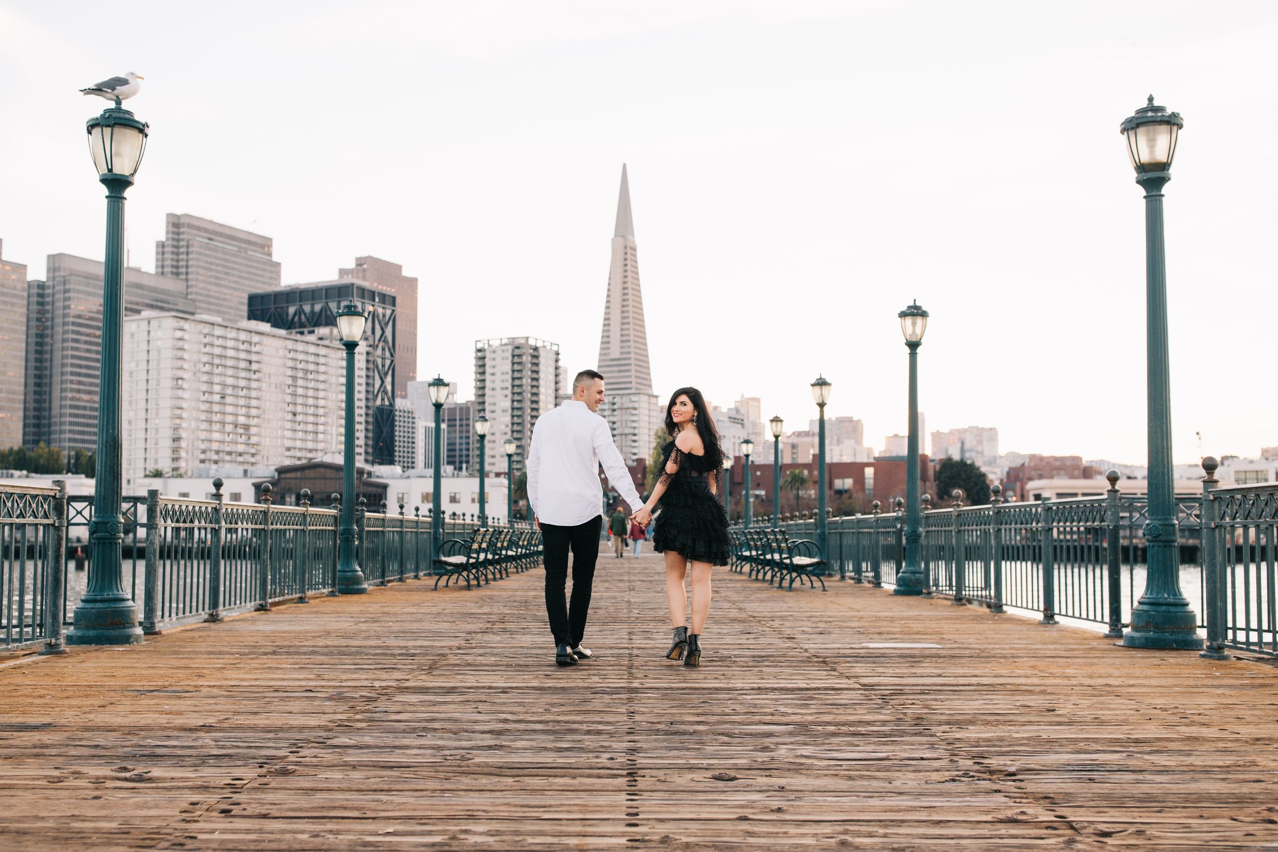A stylish couple walks along a waterfront pier with the Transamerica Pyramid tower visible in the background.