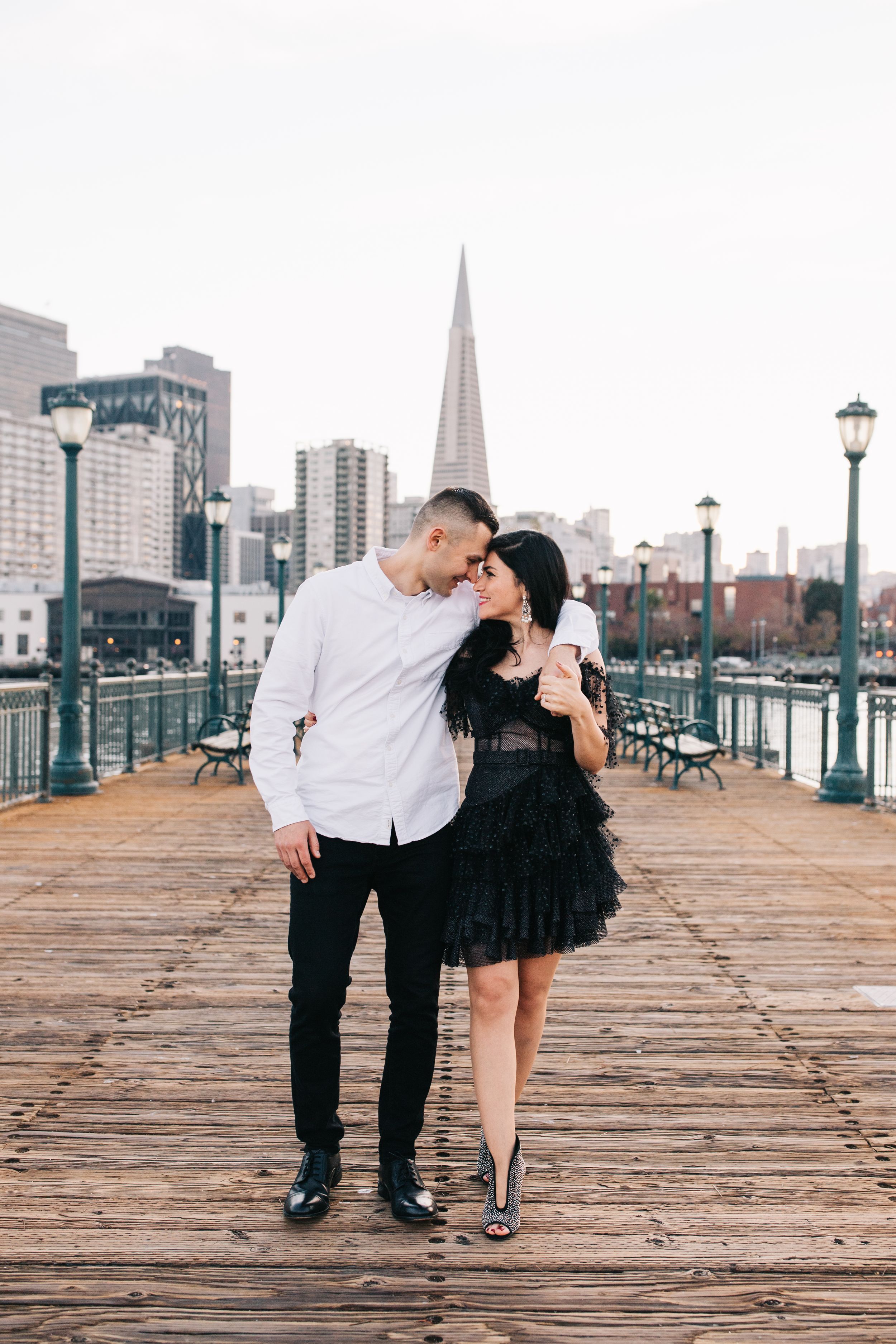 A couple in contrasting black and white outfits pose on a wooden pier with city views.