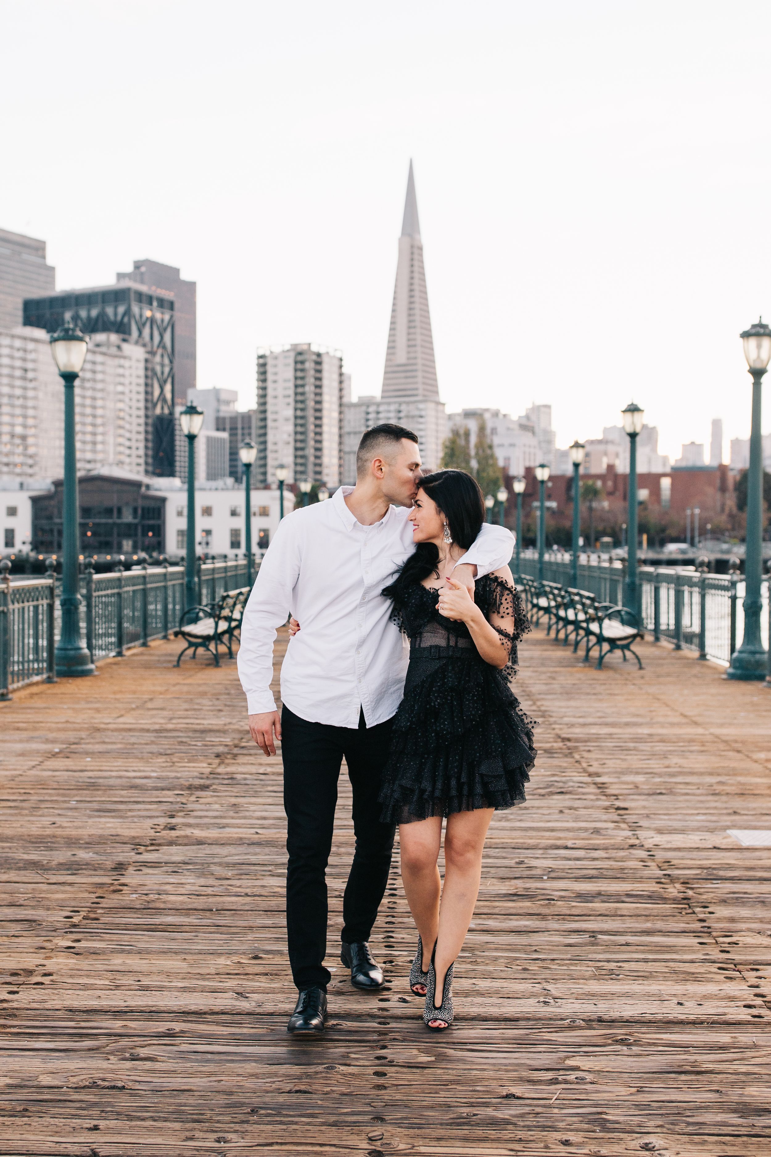 A couple shares a loving moment while walking on a pier with the San Francisco cityscape behind them.