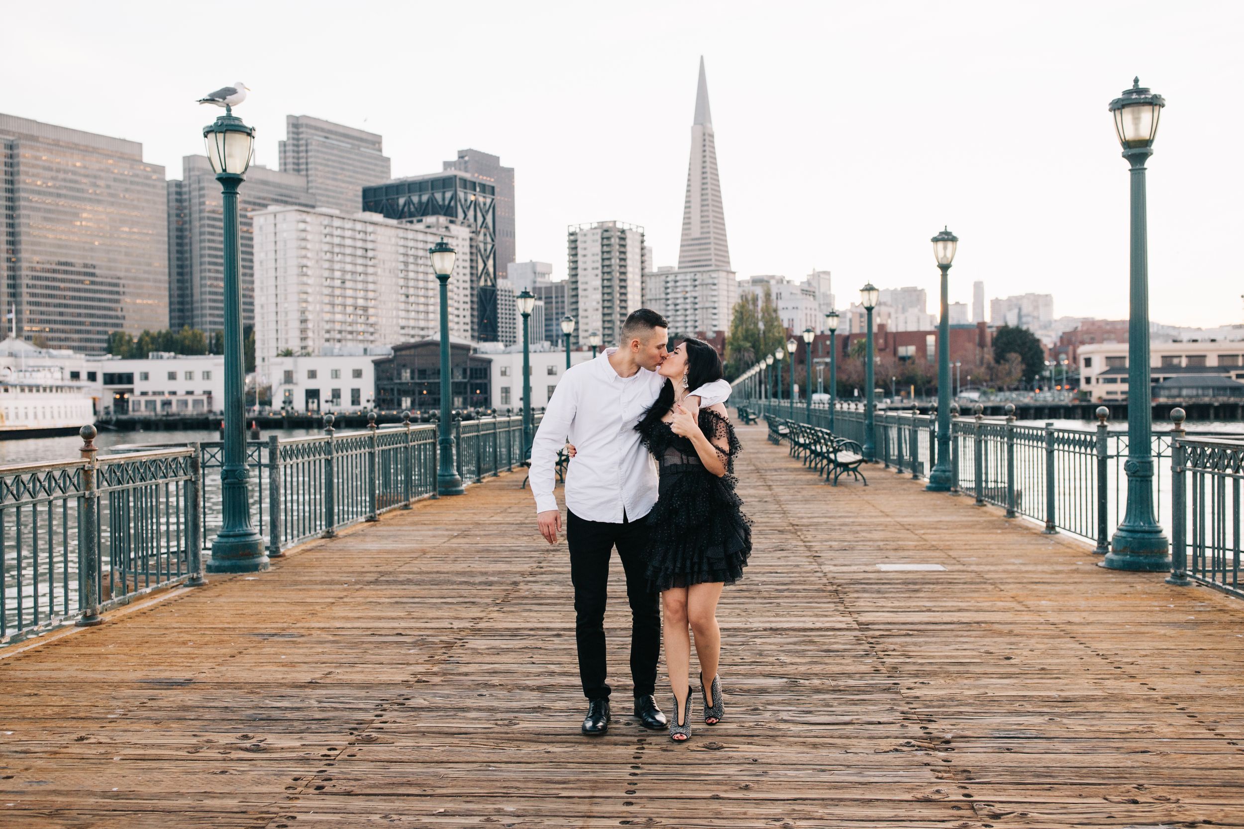 A couple embracing on a waterfront pier with San Francisco's iconic downtown buildings behind them.
