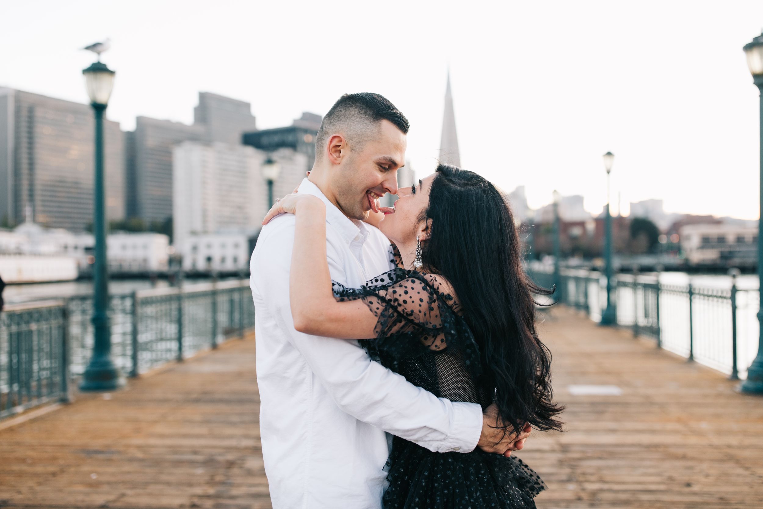 A joyful embrace on a wooden pier with urban architecture and lampposts creating a romantic setting.