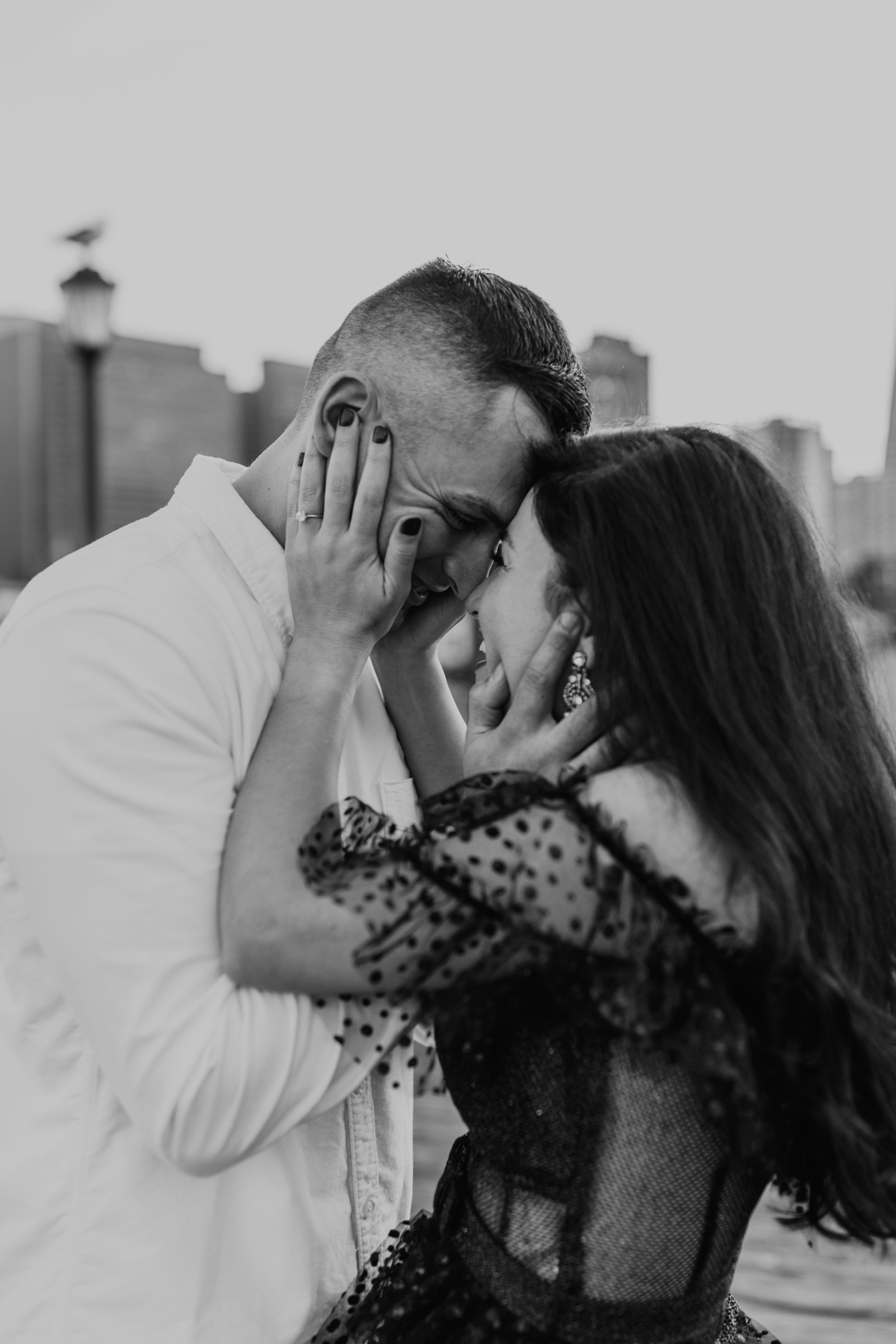 An intimate black and white portrait captured on a pier with the city skyline in soft focus.