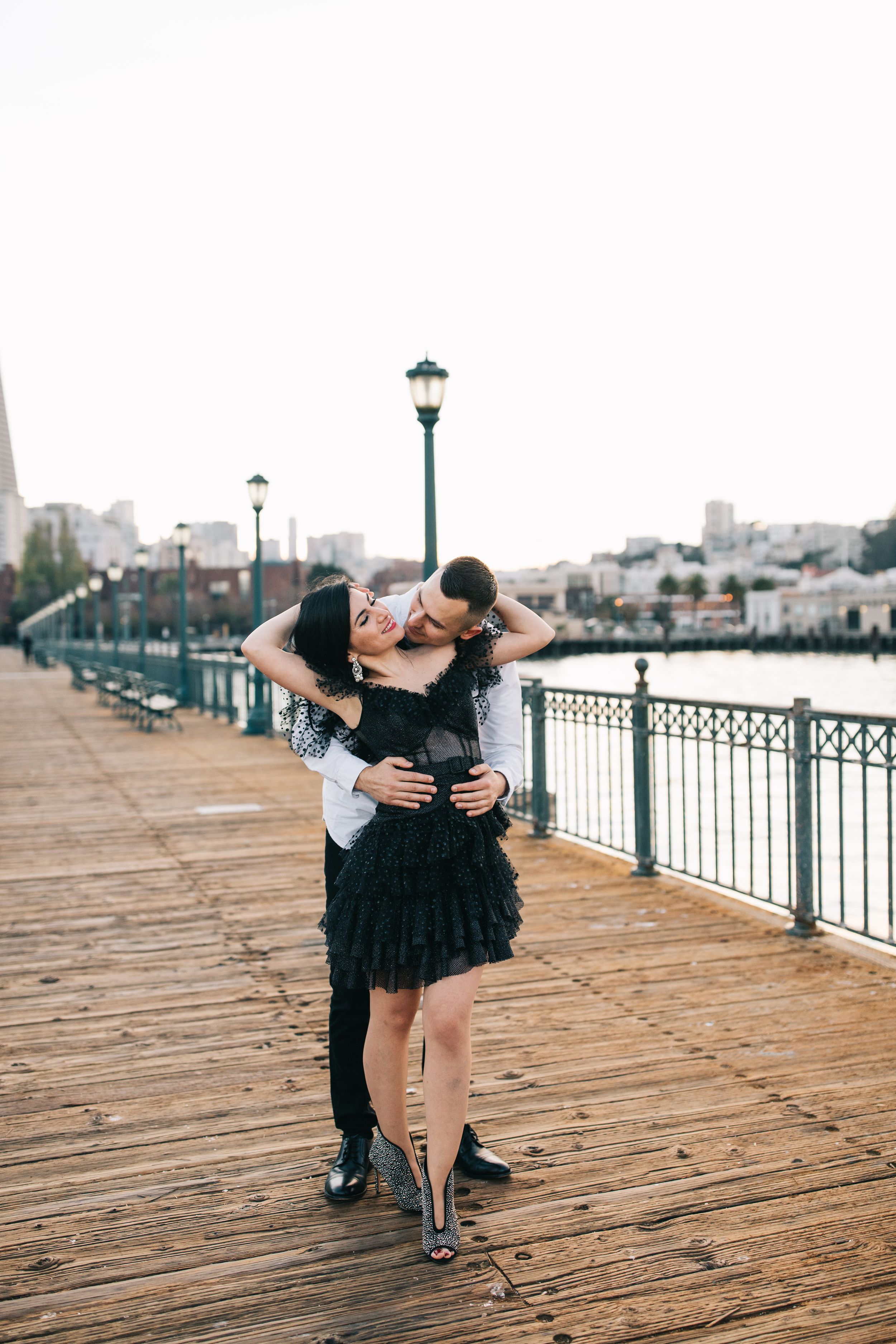 A playful dance moment captured on a wooden boardwalk with vintage-style lampposts and city views.