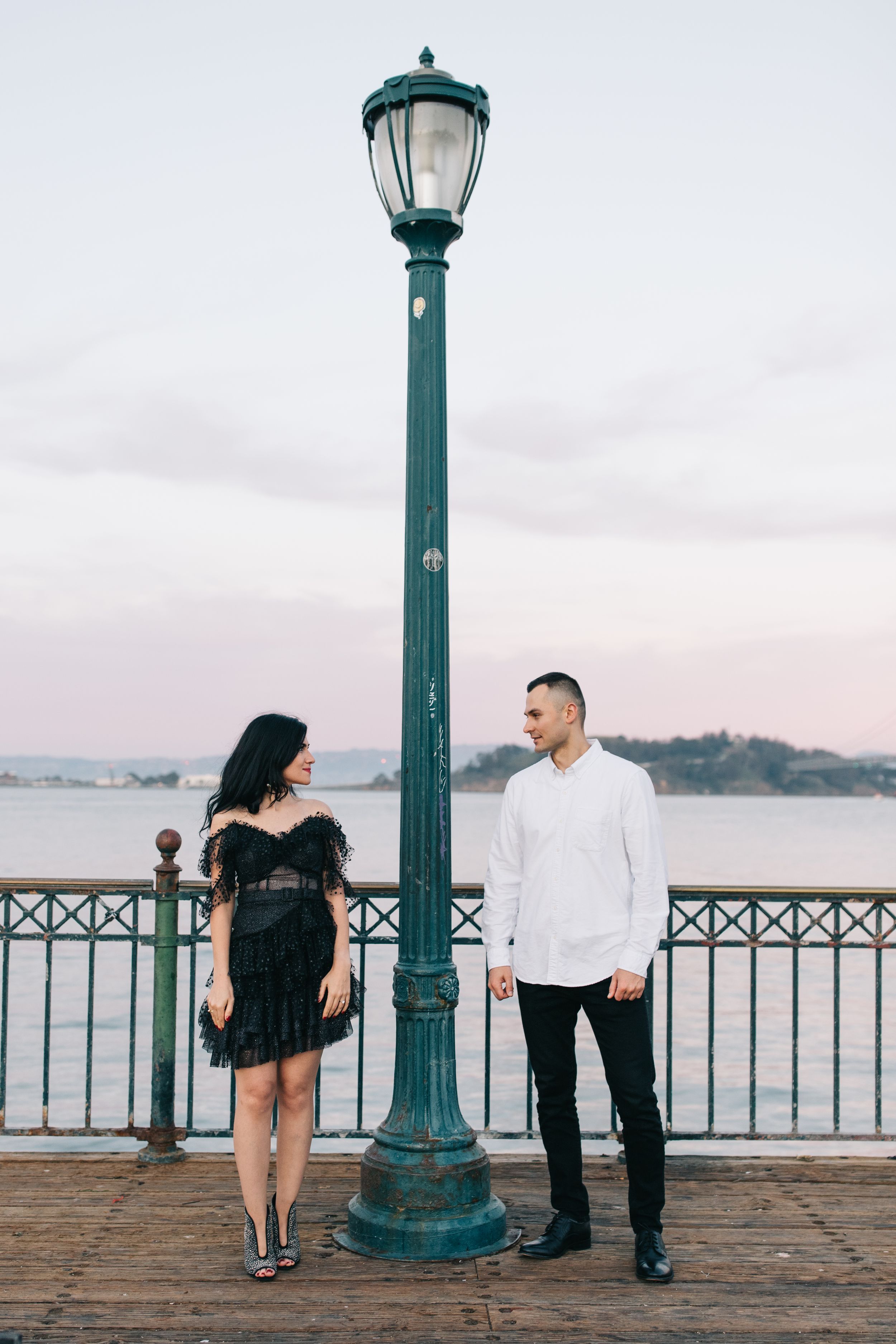Stylish couple posing by vintage lamp post on wooden pier with bay views.