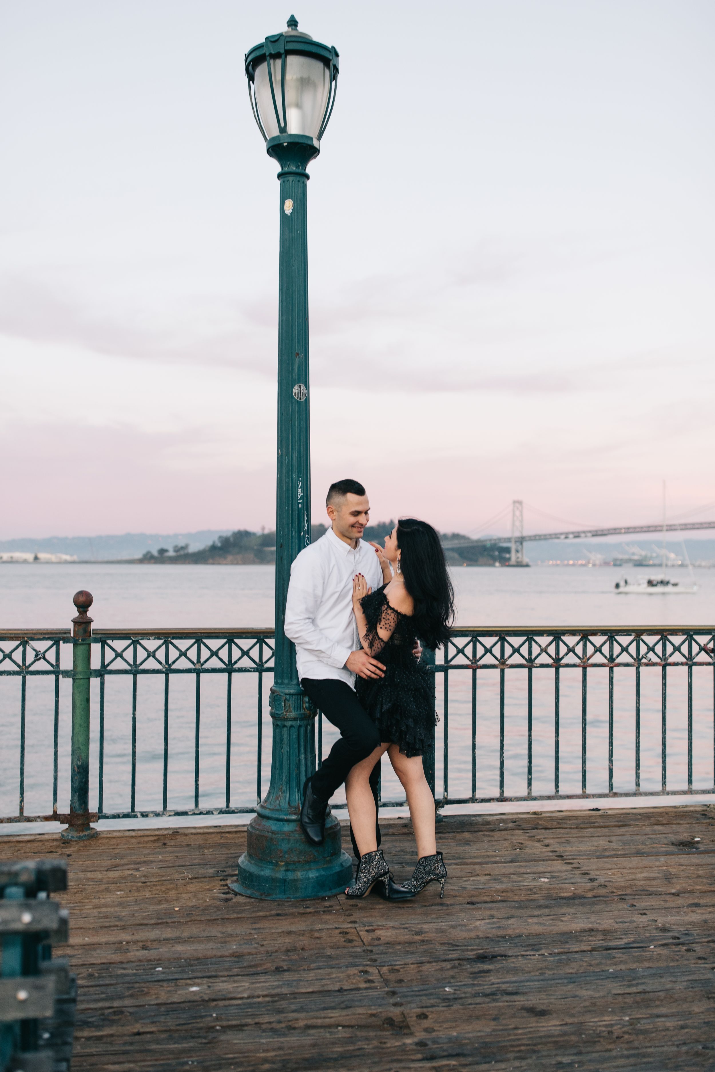 Couple embracing by teal lamp post on pier with bridge visible in background.