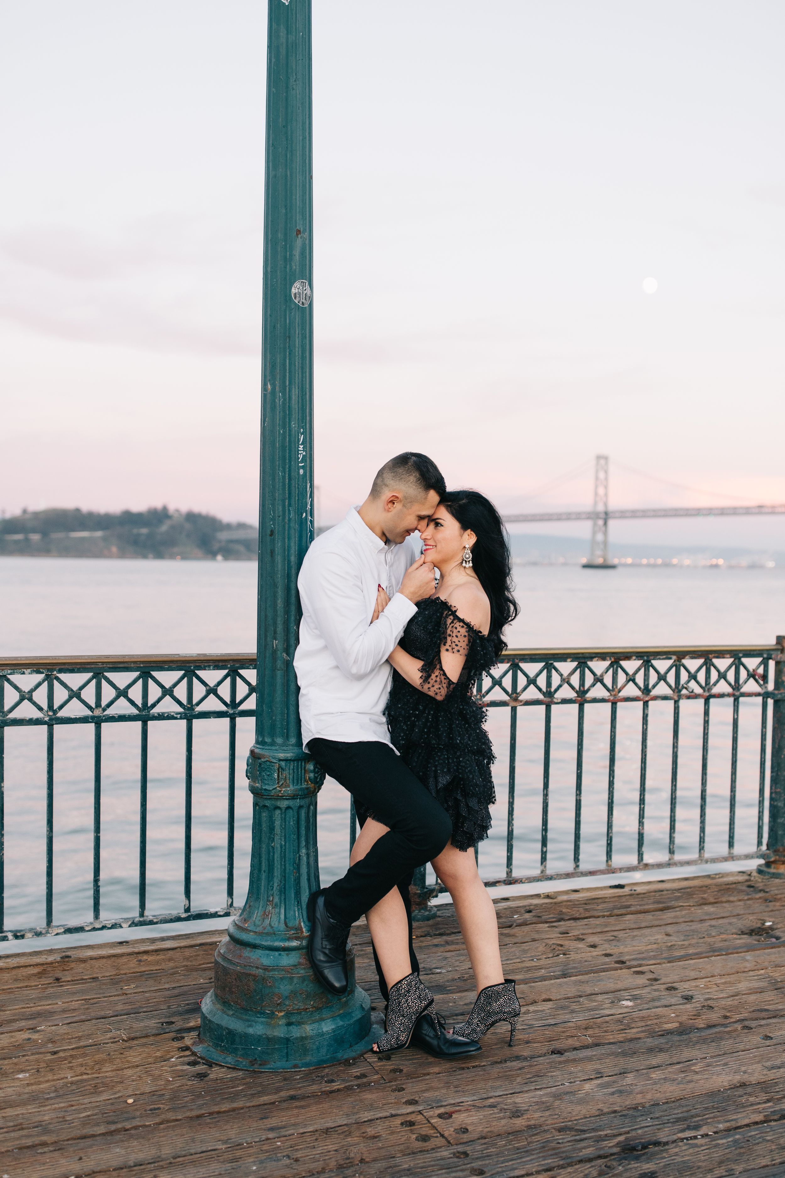 Romantic couple embracing by vintage lamp post with bay bridge in background.