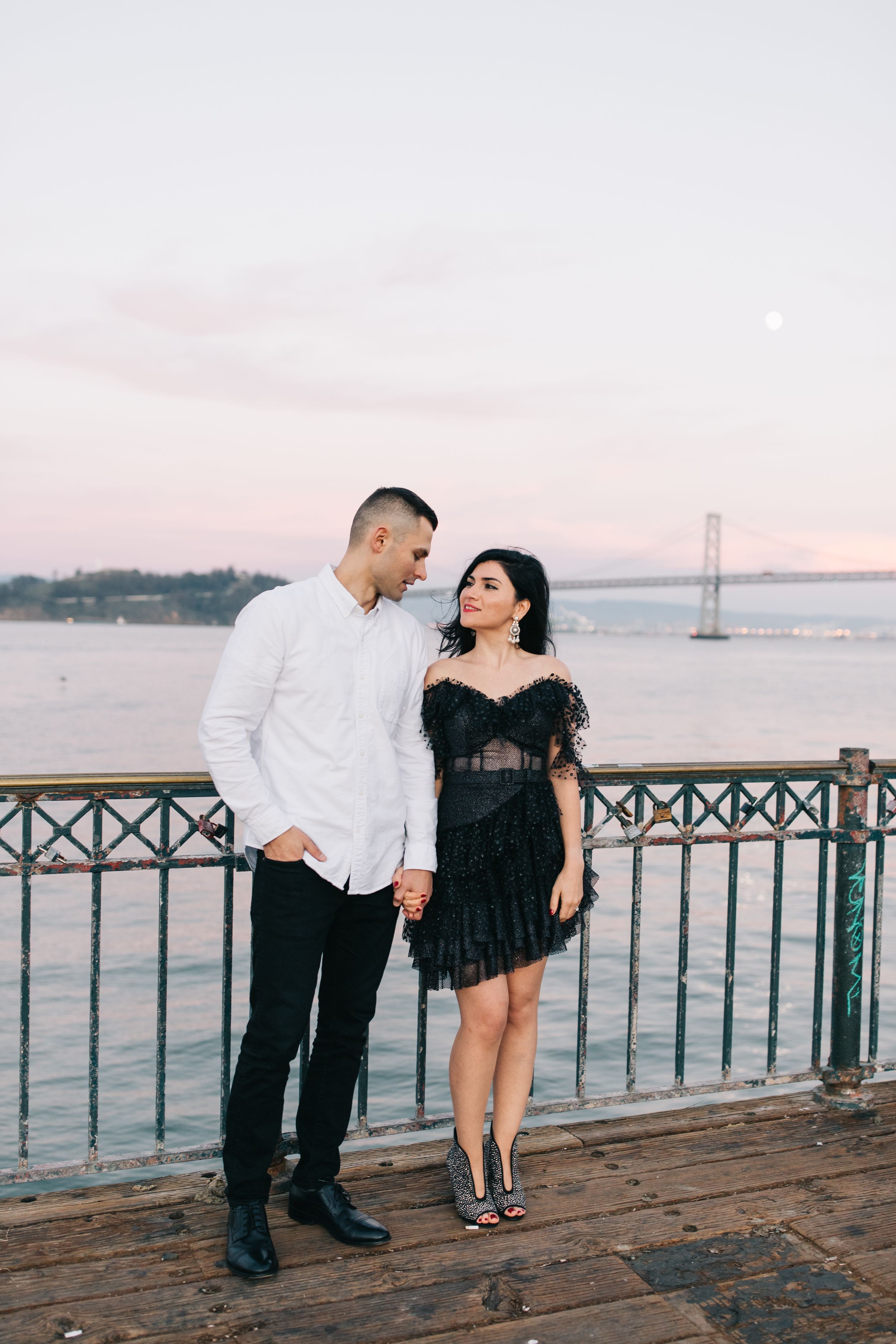 Elegant couple in formal attire standing on wooden pier at sunset.