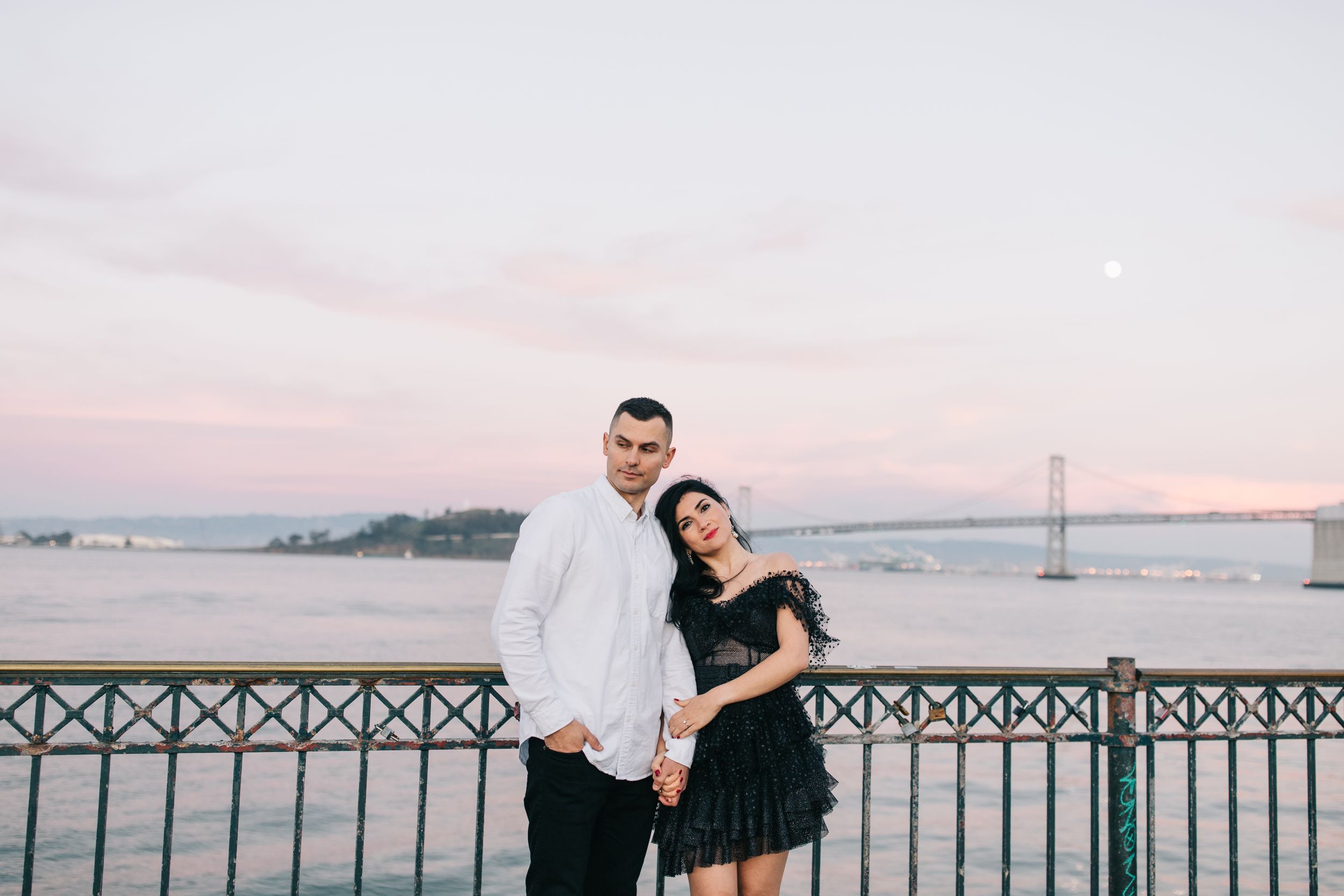 Happy couple posing on pier with San Francisco Bay Bridge visible in distance.