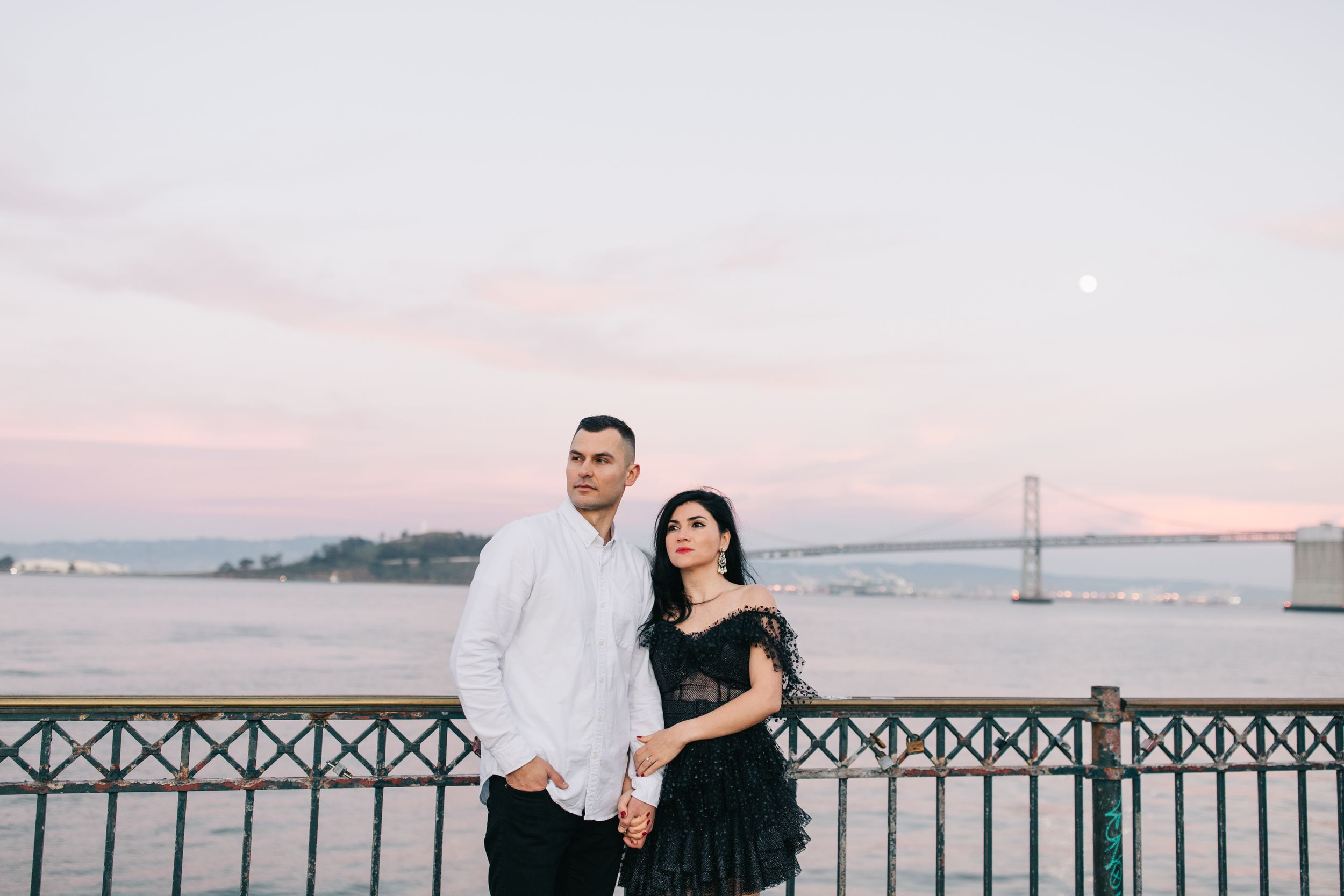 Couple in coordinated outfits pose against waterfront backdrop during golden hour.