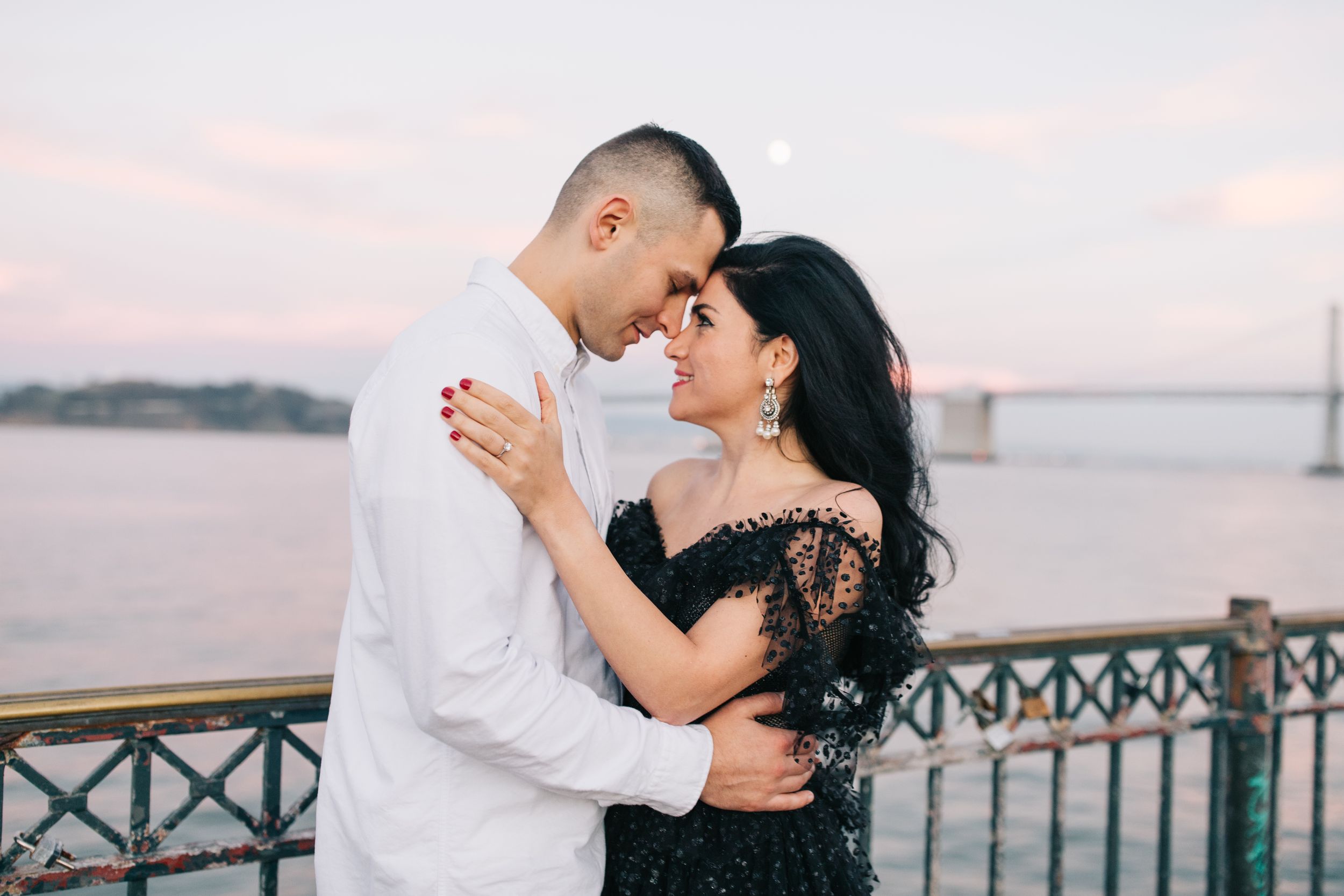 Romantic couple embracing on a pier during sunset with a bridge visible in the distance.