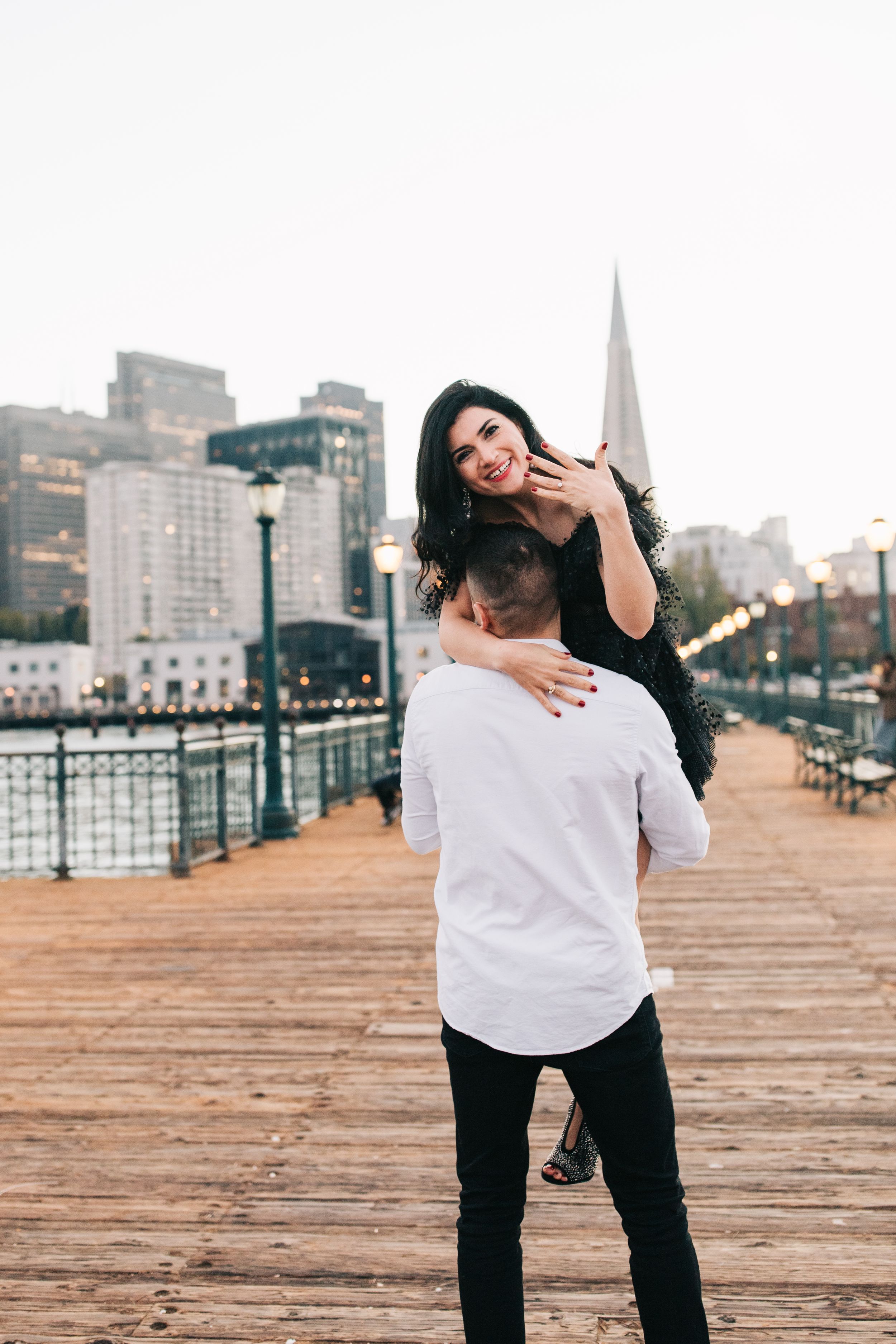 Couple sharing playful moment on San Francisco pier with downtown skyline in background.