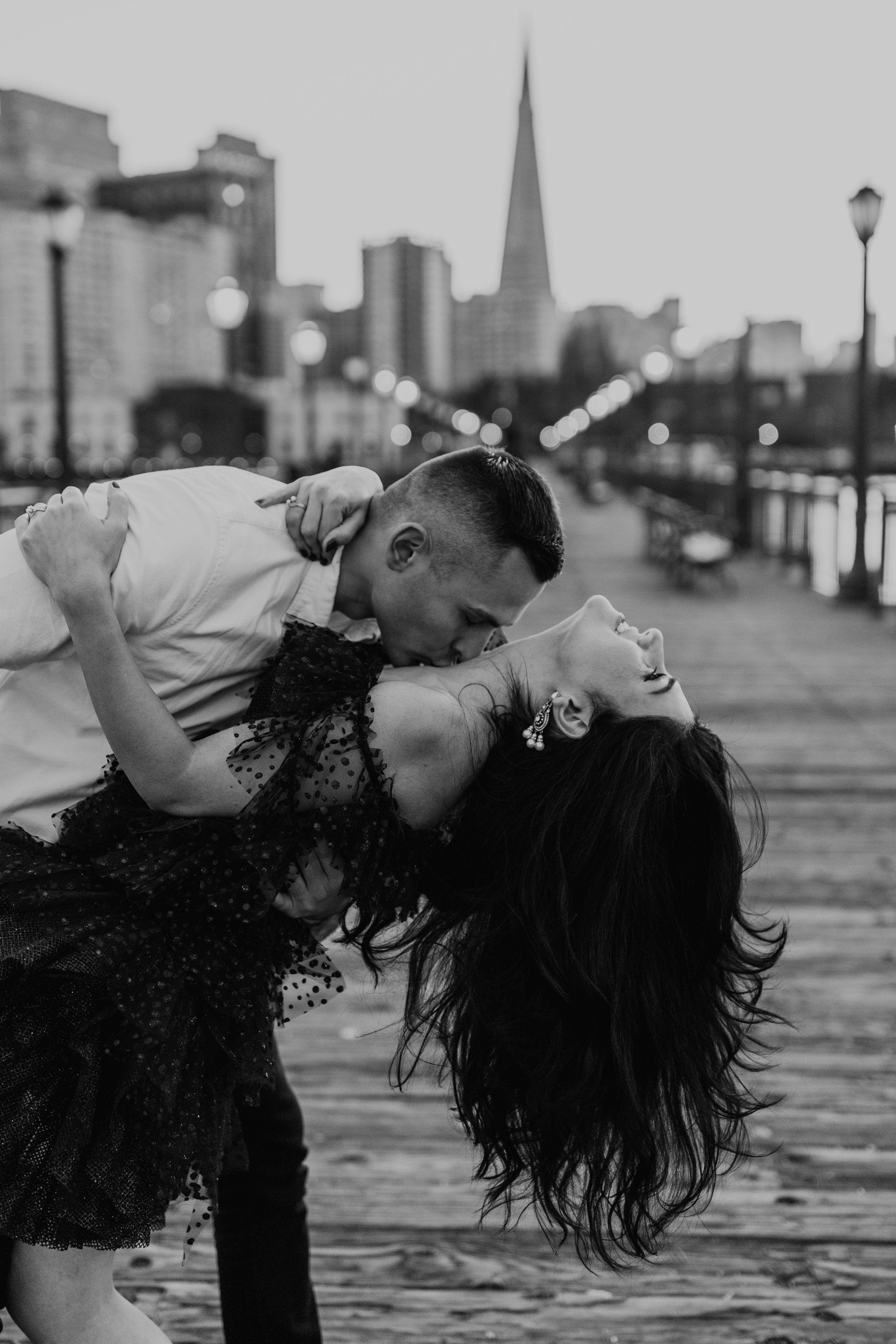 Dramatic black and white photo of couple dancing on pier with city skyline.