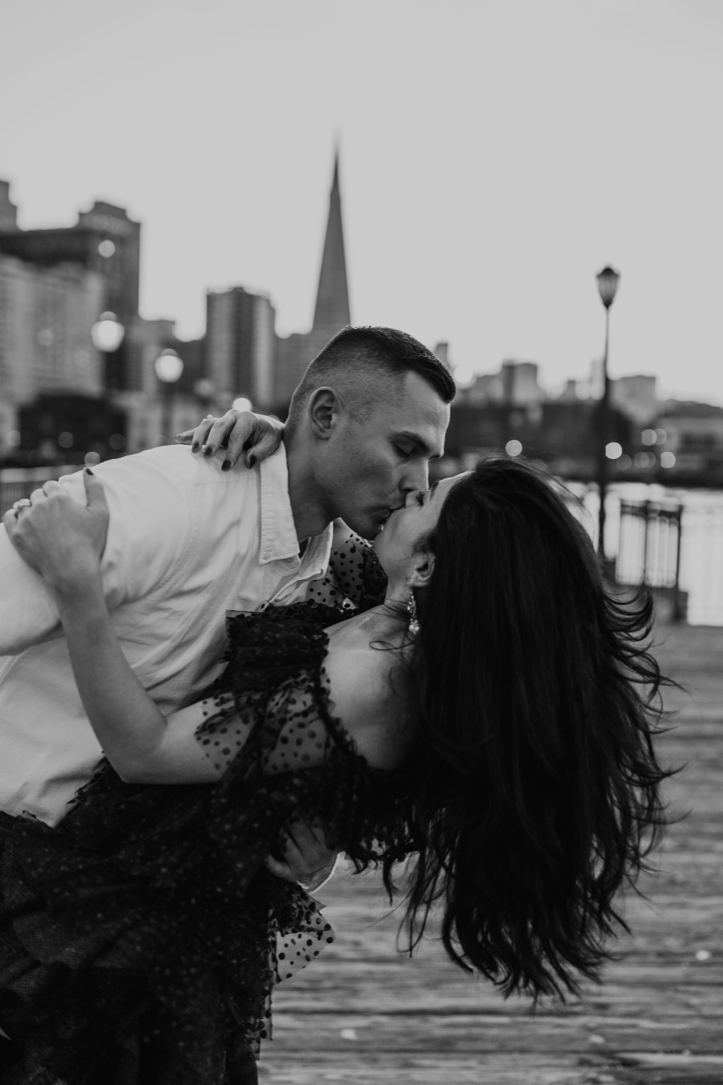 Romantic couple dips on wooden pier with city skyline in black and white.