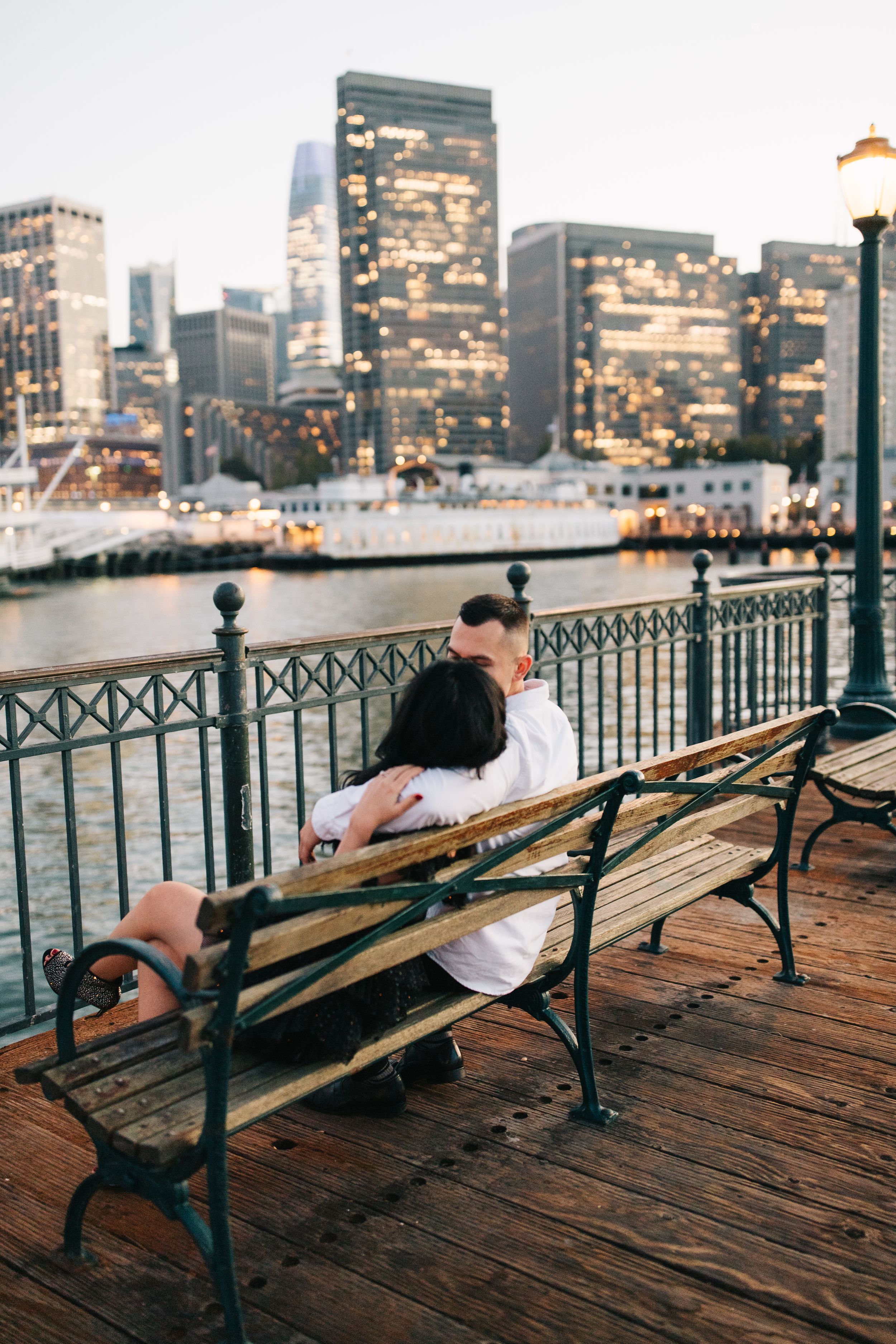 Couple resting on bench overlooking illuminated city skyline and harbor at dusk.