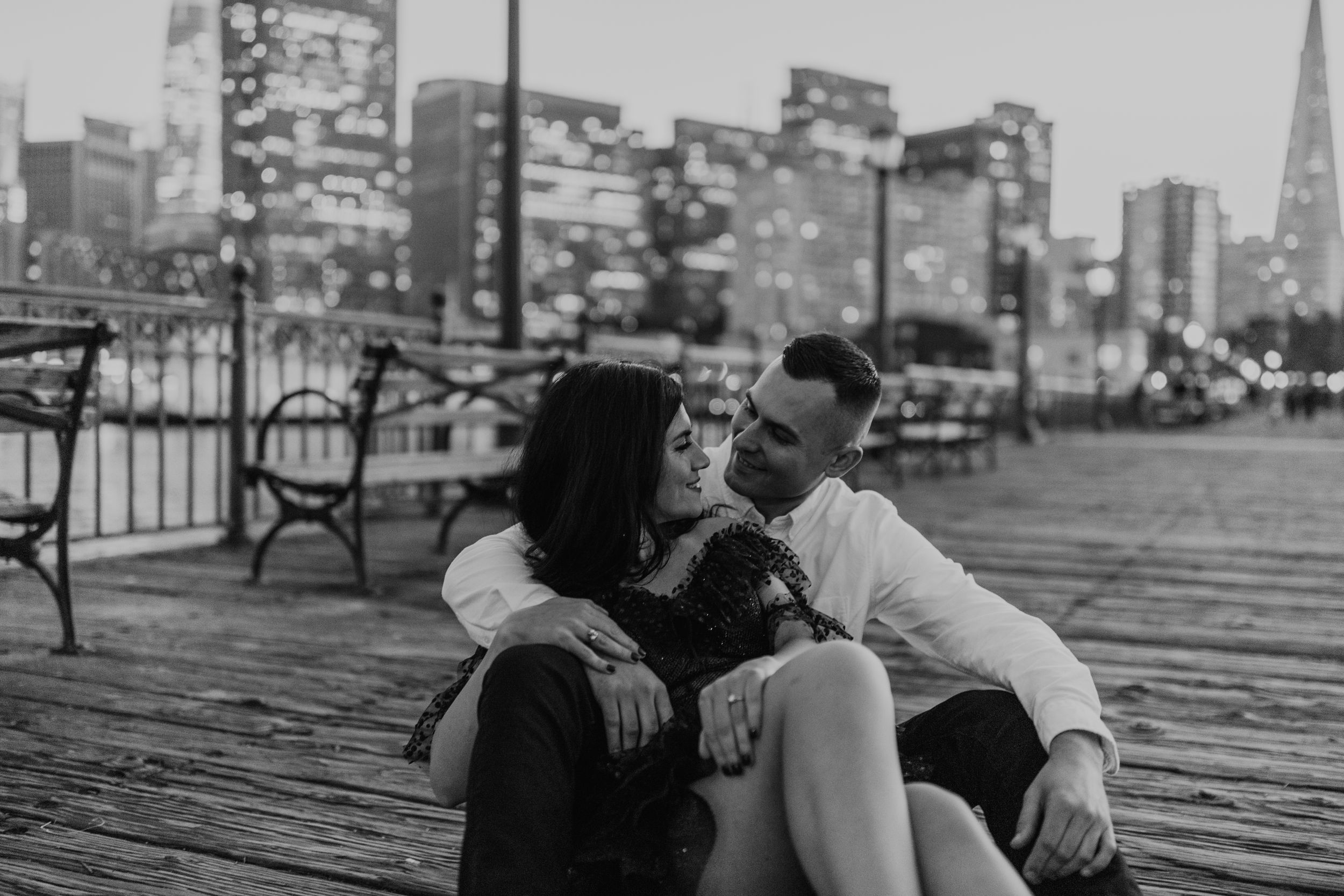 Black and white photo of couple sharing intimate moment on pier with urban skyline backdrop.