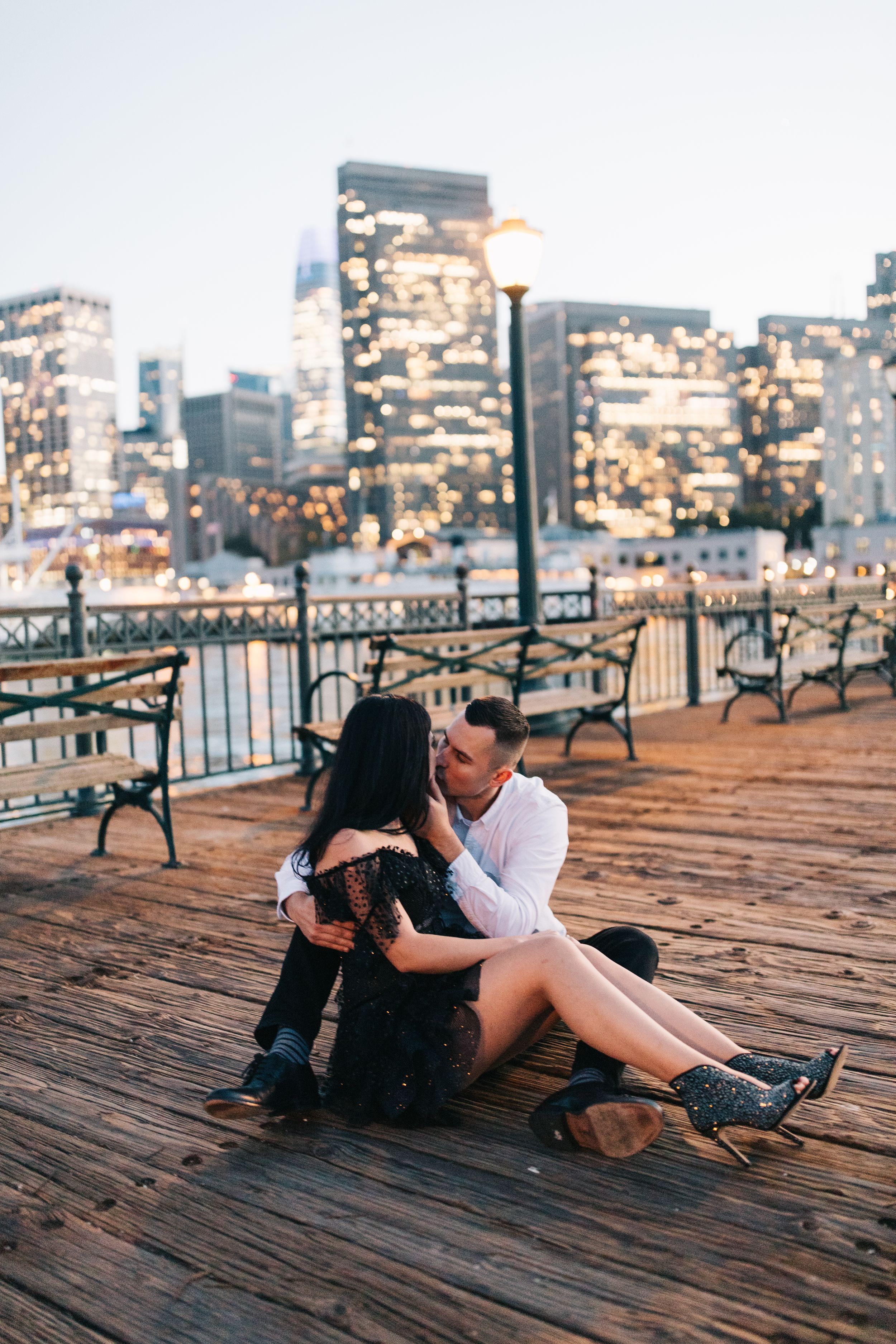 Romantic sunset moment on wooden pier with glowing city buildings and lampposts in background.