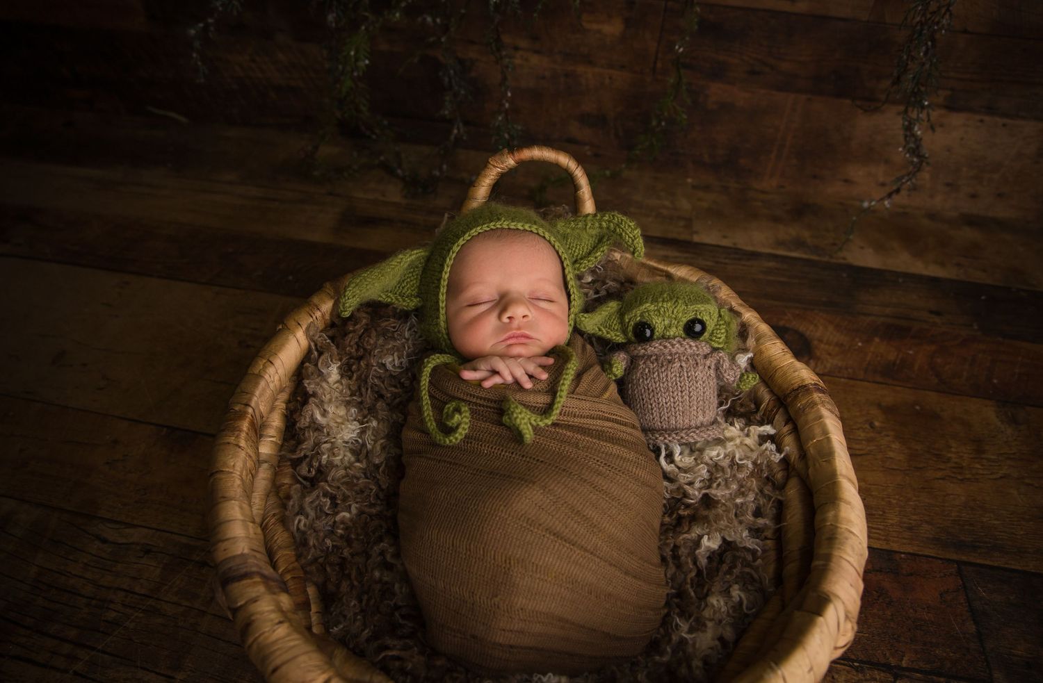 Newborn in Yoda-themed outfit sleeping in rustic wooden basket with plush toy.