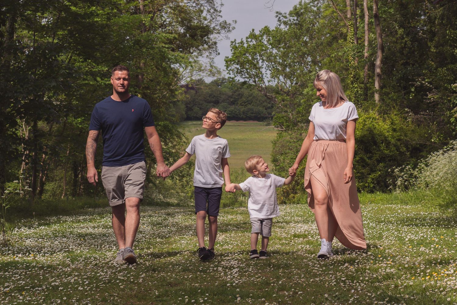 A young family all holding hands walking towards the camera. Parents are on the ends and boys in the middle. Each boy is looking at a parent