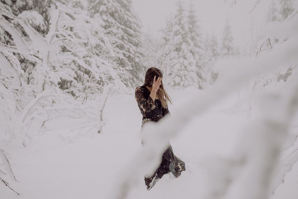 A person in winter clothing walks through deep snow surrounded by frost-covered pine trees.