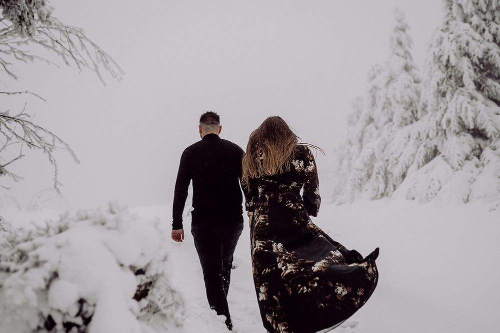 A couple walks away through a snowy forest path with snow-laden evergreen trees.