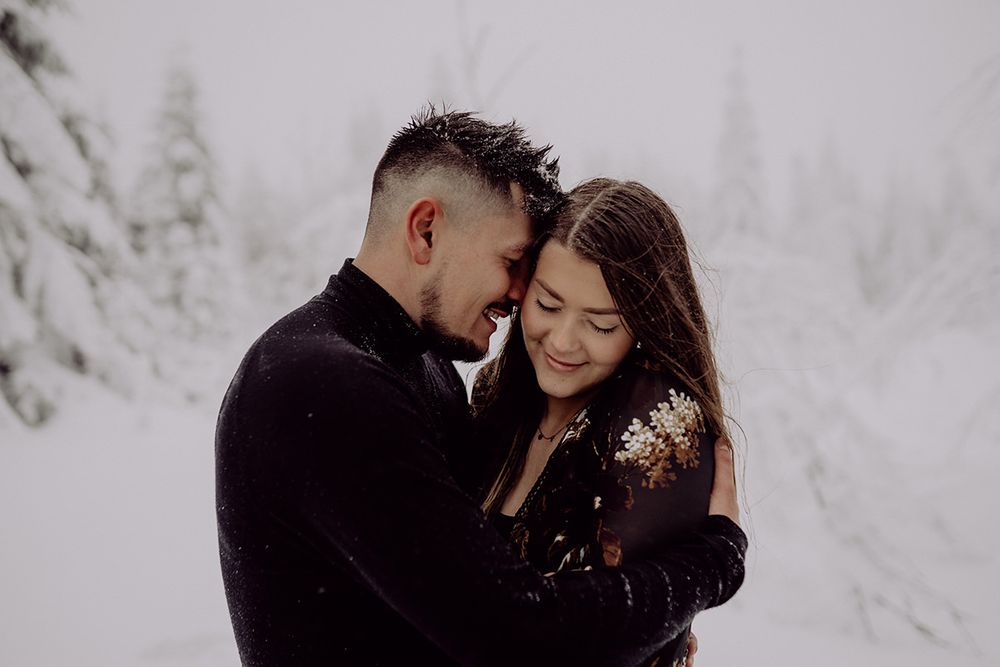 A romantic couple embraces in a snowy winter forest setting with snow-covered trees in the background.
