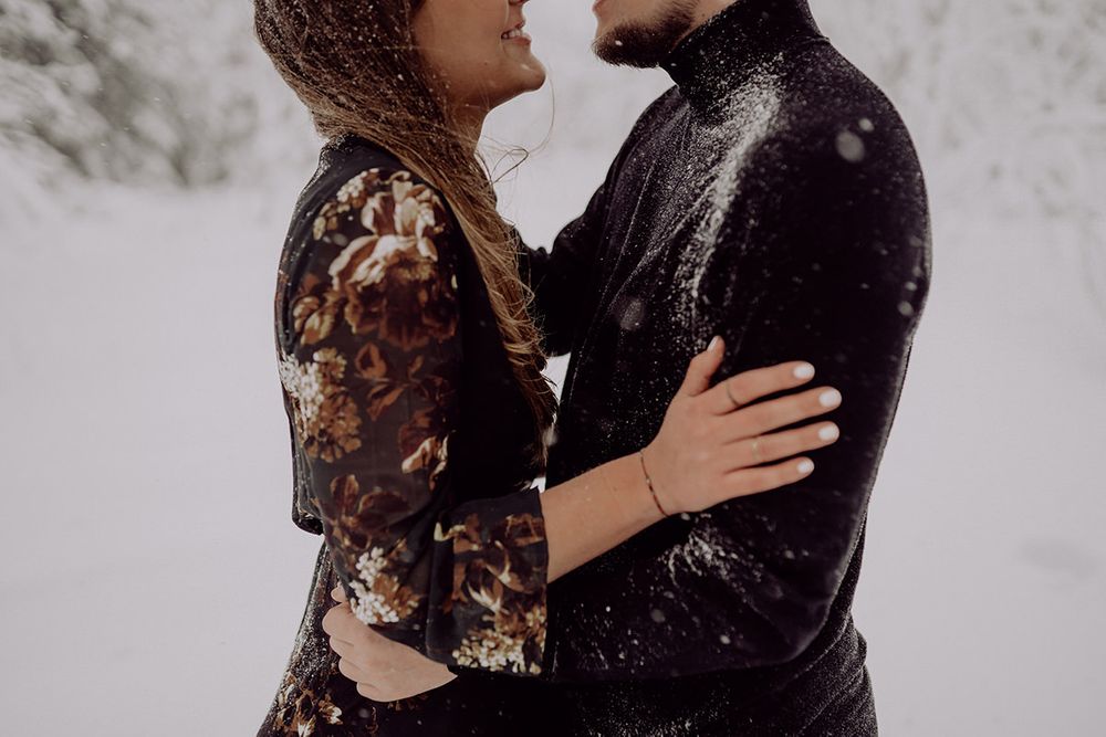 A couple embraces in falling snow surrounded by winter forest scenery.