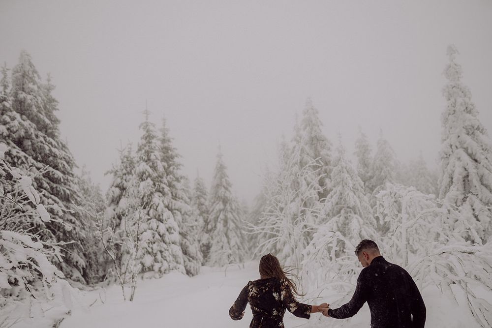 A couple holds hands while walking through a snow-covered forest of evergreen trees.