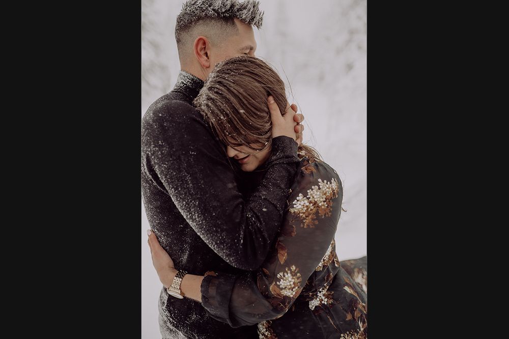 A couple embraces tenderly in winter clothing against a snowy backdrop.