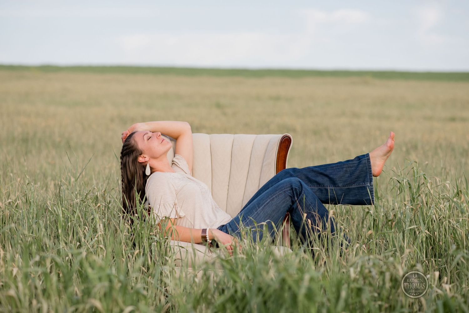 Someone reclines playfully in a beige armchair placed in a grassy field.