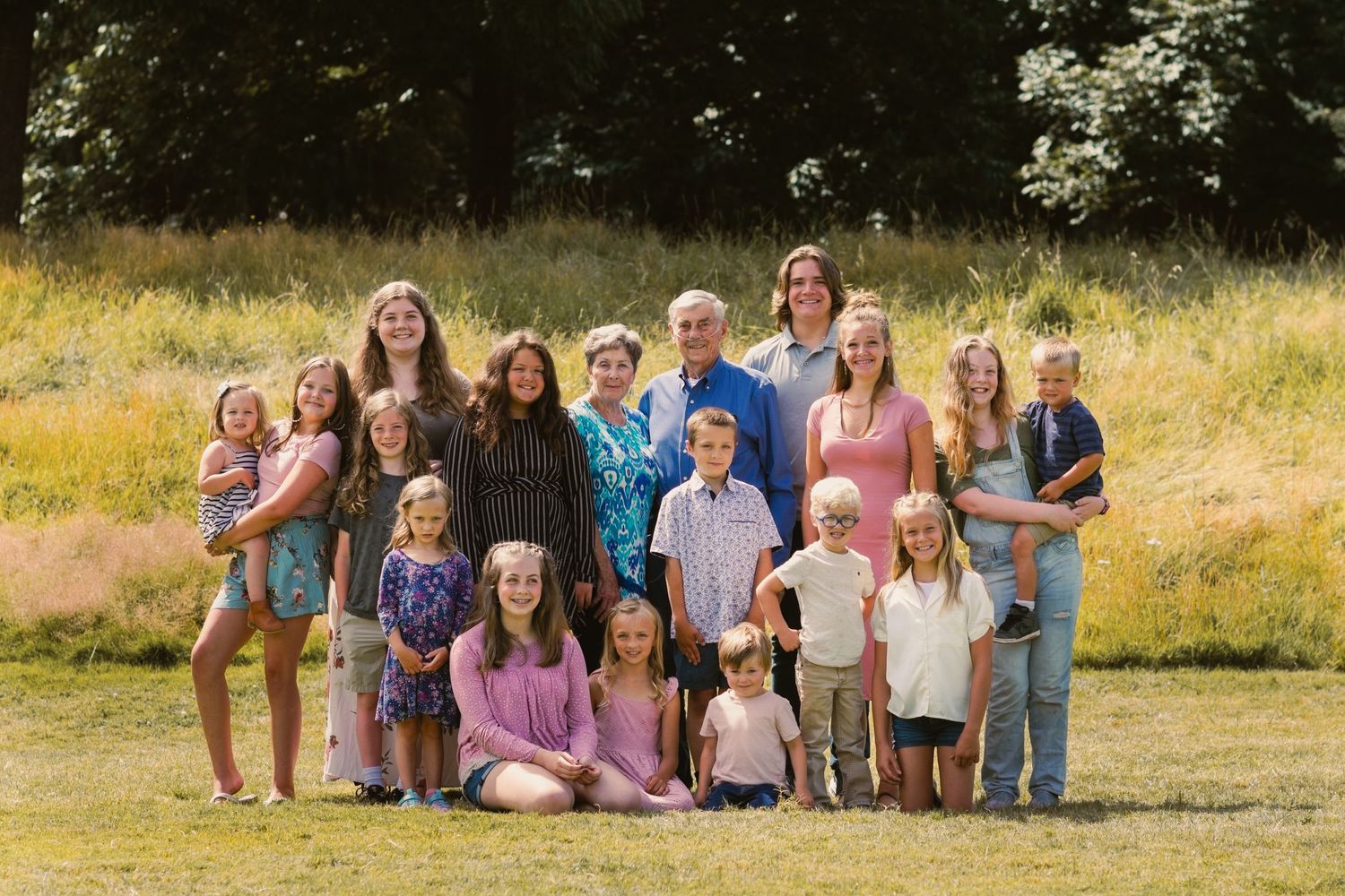 family of 15 grandkids posing for camera with great grandparents in a field on a sunny day with trees in the background