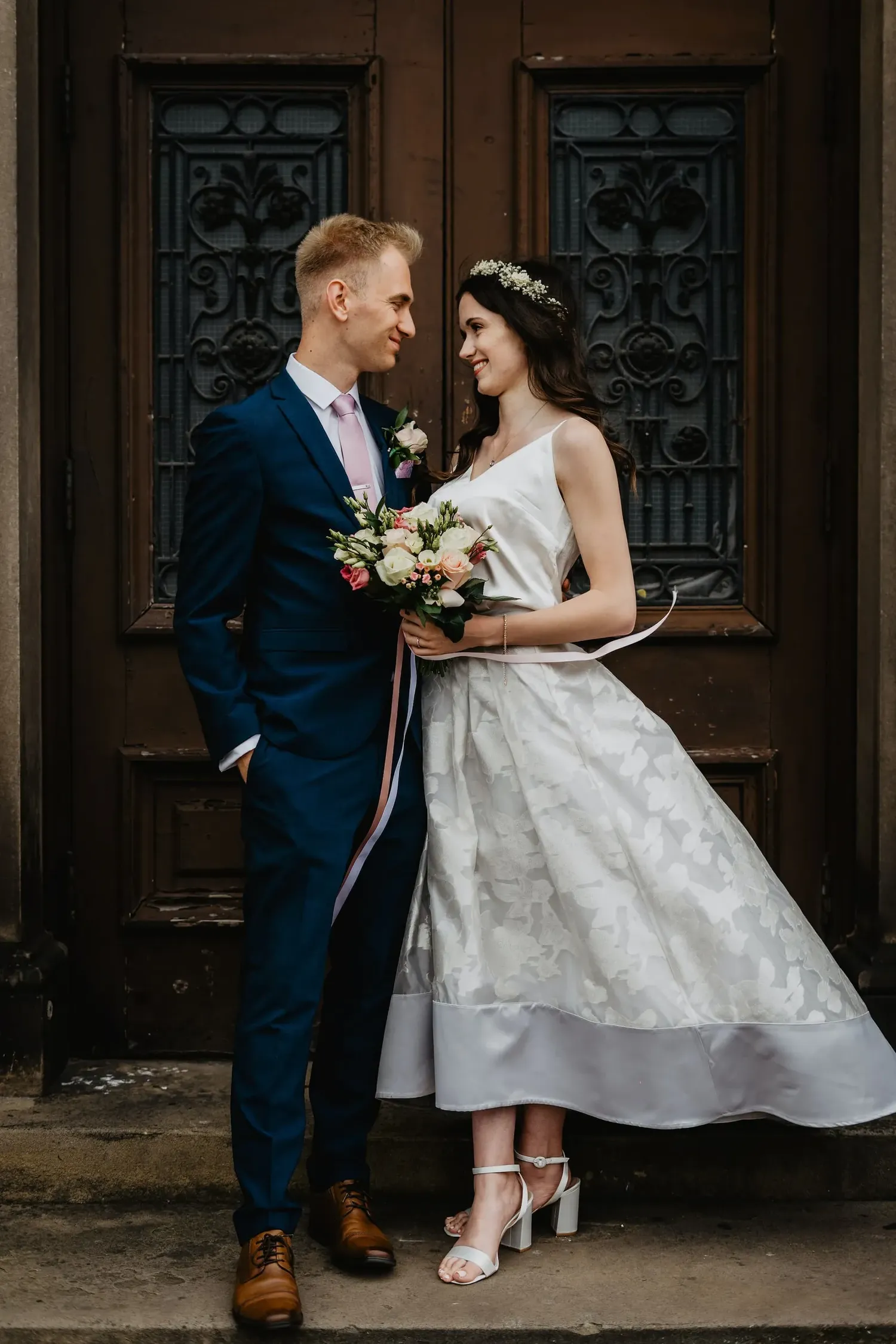 A romantic wedding portrait against ornate brown doors with bride in tea length gown and groom in navy suit.