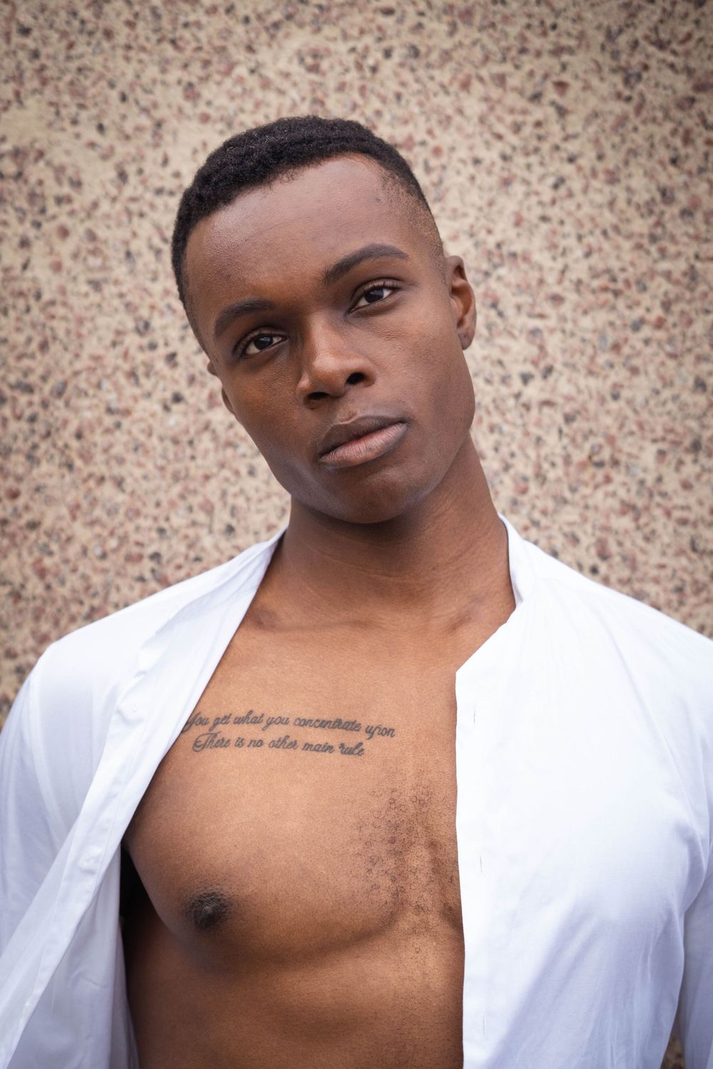 Black Athletic person in open white shirt shows chest tattoo against textured wall.