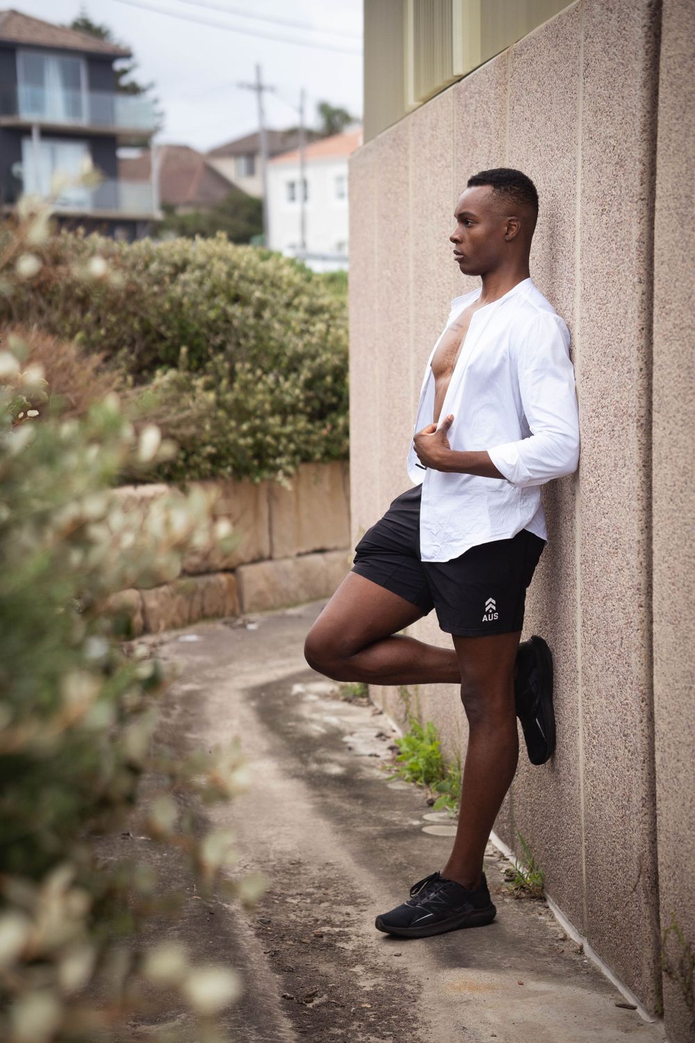 Black Athletic person in white shirt and black shorts leans against beige wall outdoors.