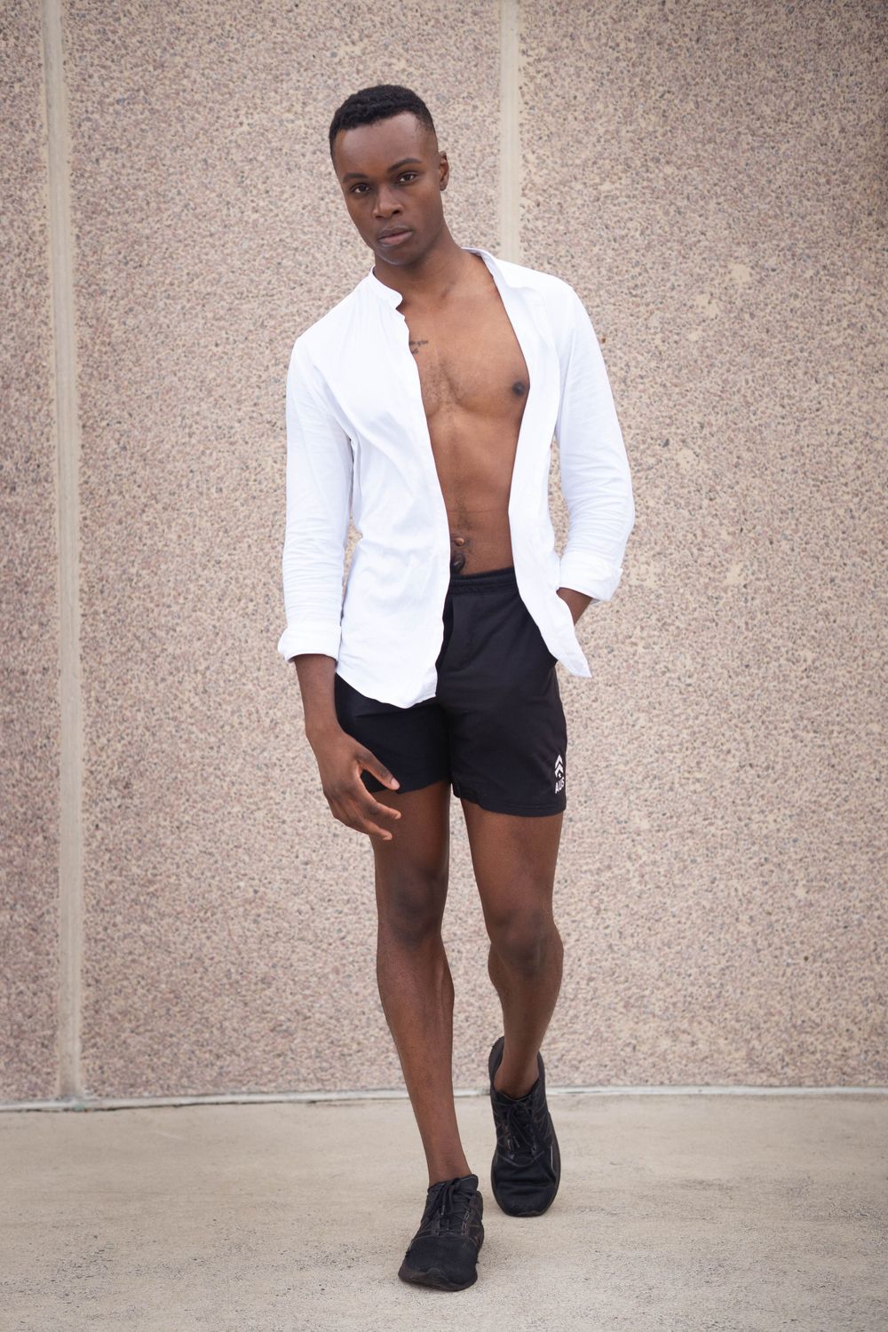 Black Athletic model poses in white open shirt and black shorts against textured wall background.