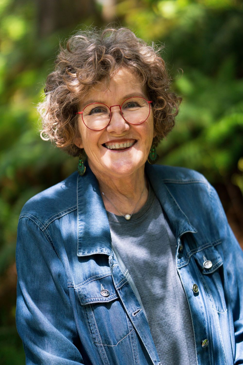 Older lady with curly hair and glasses smiling while wearing denim jacket in garden setting.