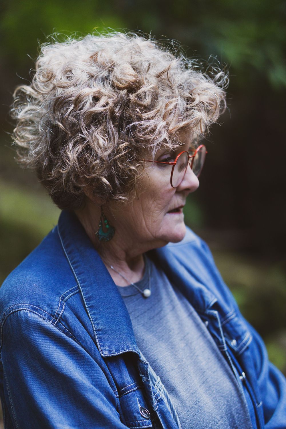 Older lady with curly grey hair and glasses wearing denim jacket in outdoor setting.