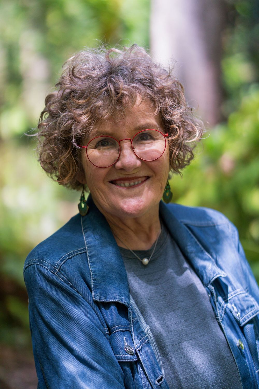 Older lady with curly grey hair and round glasses wearing denim jacket outdoors.
