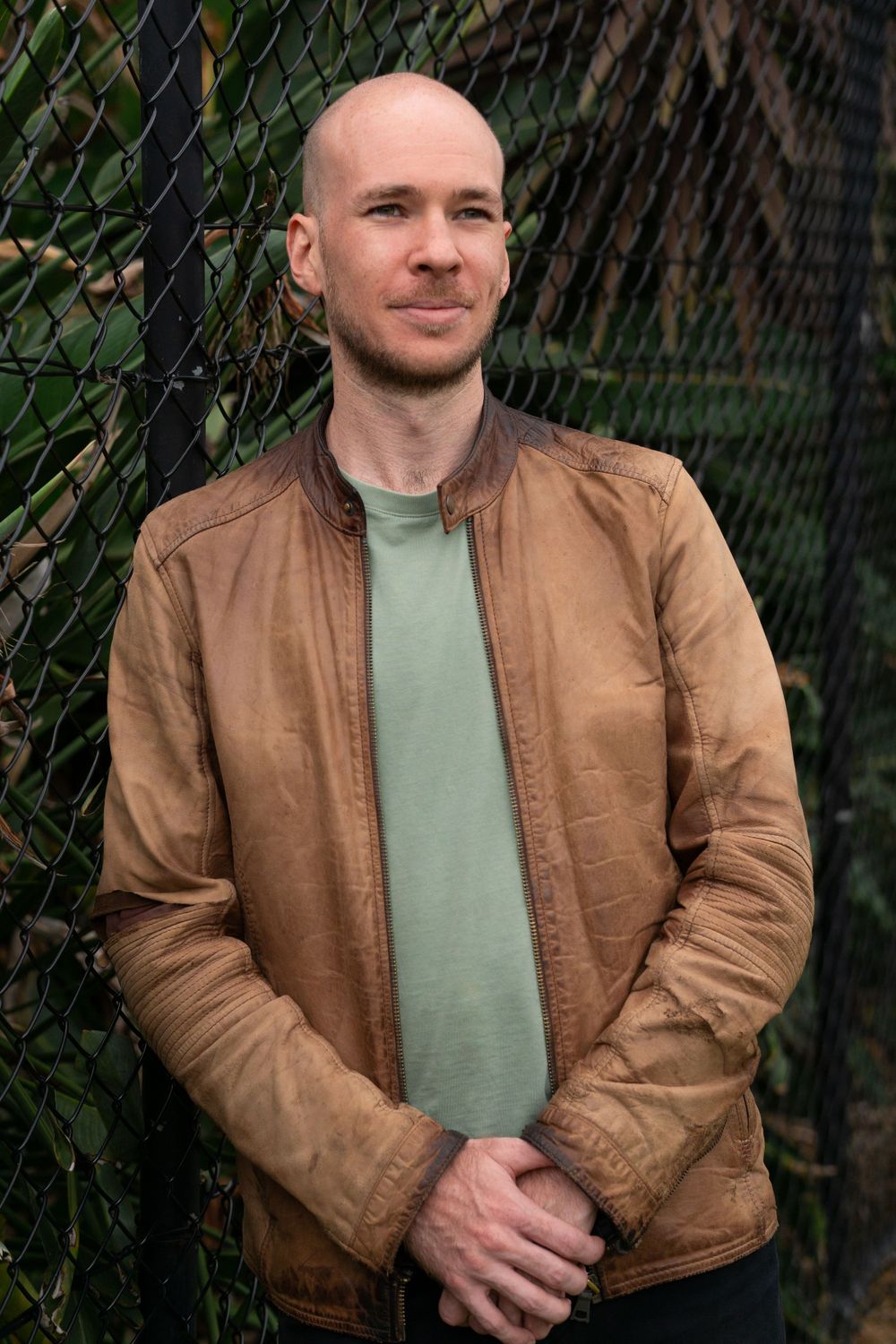 Casual portrait against chain link fence wearing brown leather jacket and sage shirt.