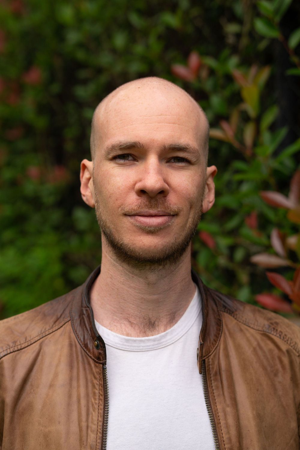Professional headshot of person in brown leather jacket against green garden background.