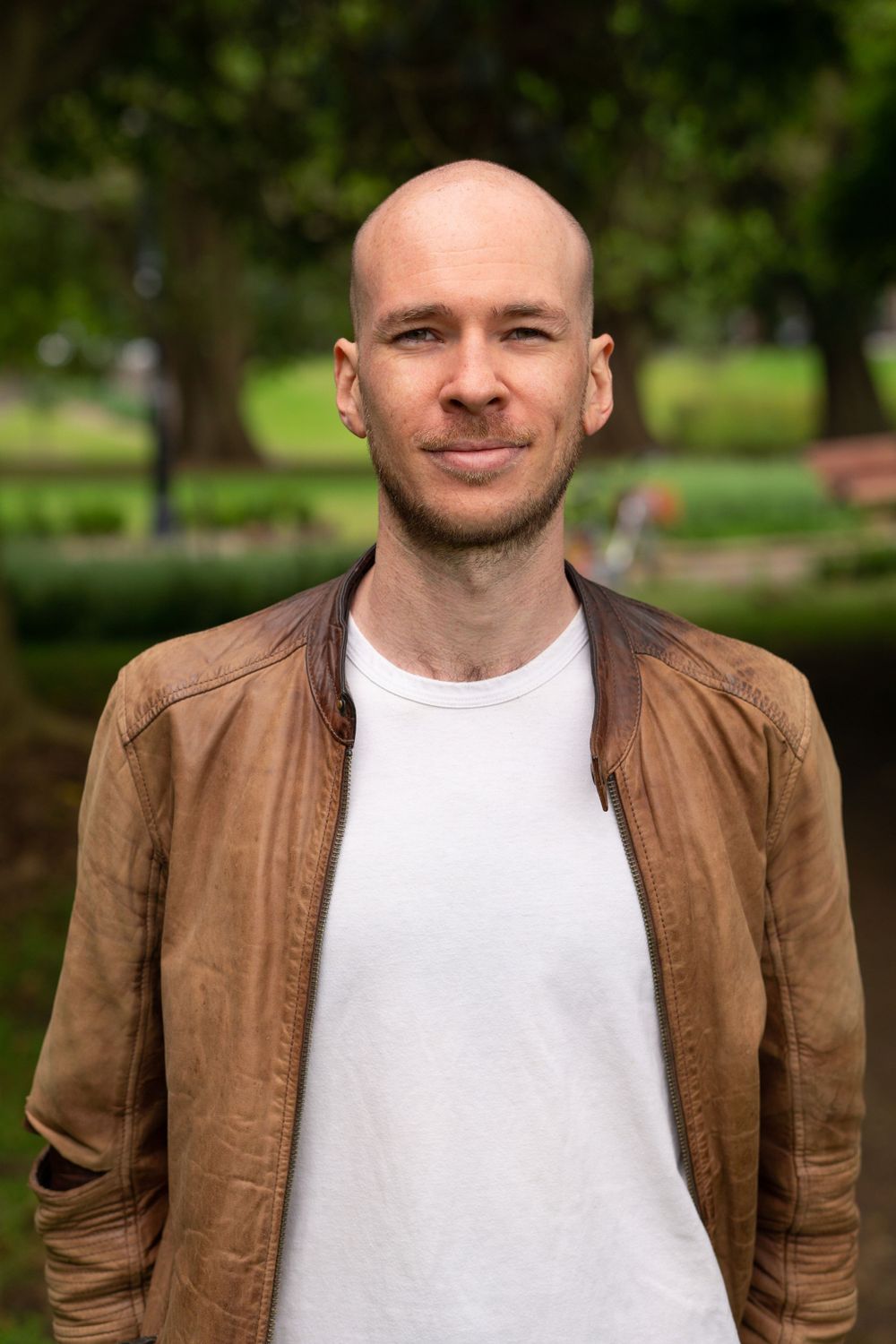 Person wearing brown leather jacket smiling at camera in outdoor setting.