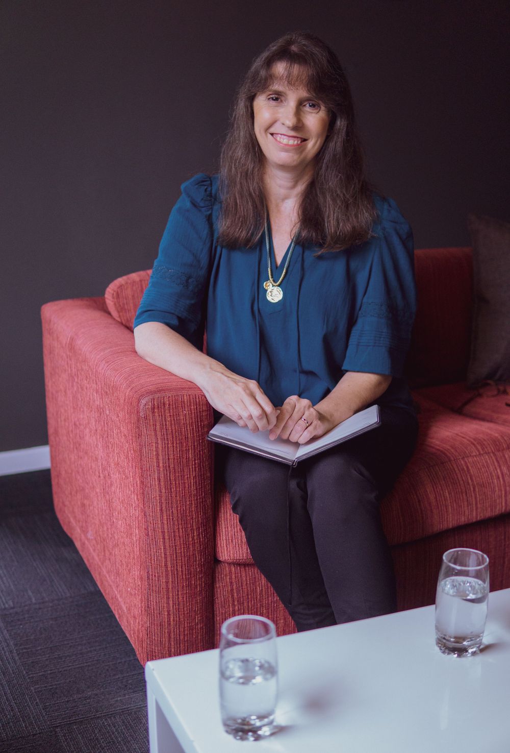 Older professional lady in blue outfit seated on red couch with notebook and water glasses on table.