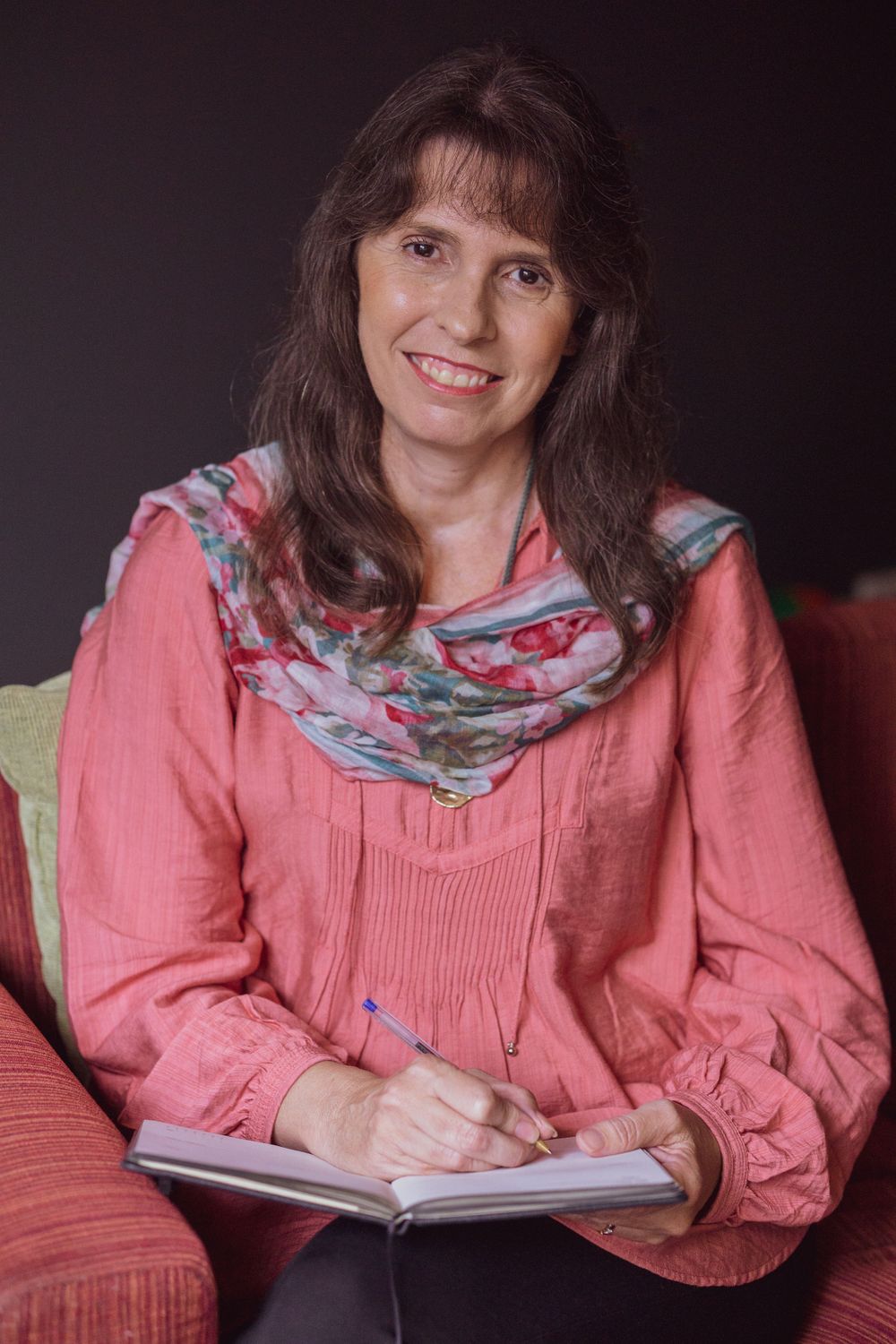 Older professional lady in pink blouse with floral scarf sitting on red couch with notebook.