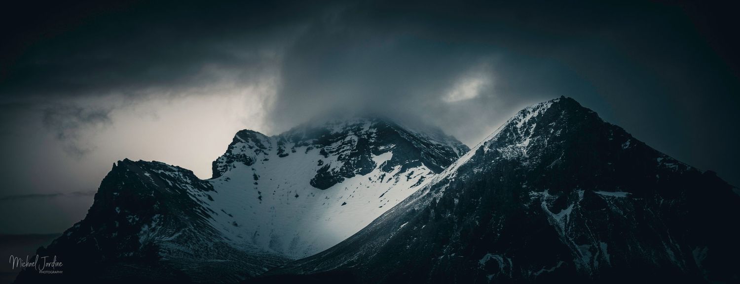 Dramatic mountain peaks under dark stormy clouds in moody panoramic landscape.