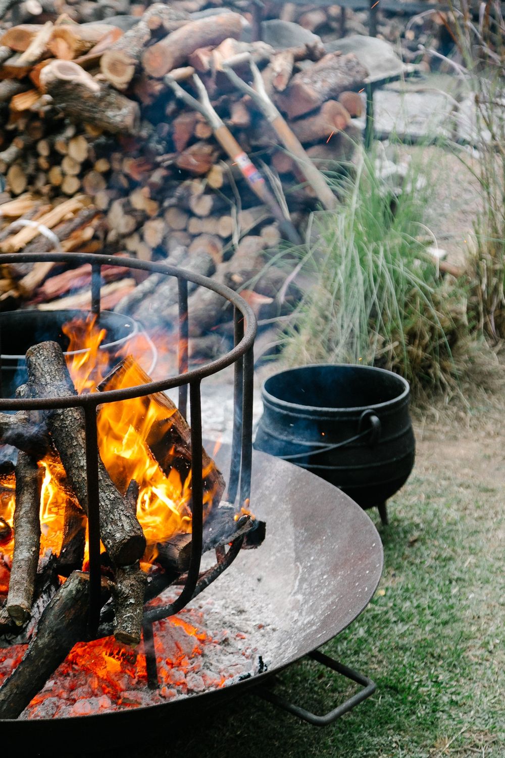 Outdoor fire pit with burning logs and flames in a metal ring near a black pot.