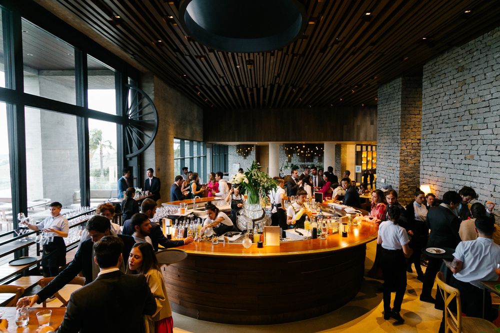 A busy restaurant interior with a curved wooden bar and diners seated throughout the modern industrial space.