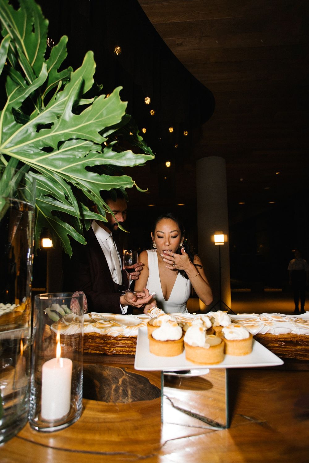 Elegant table setting with candlelight, desserts, and a large tropical leaf arrangement.