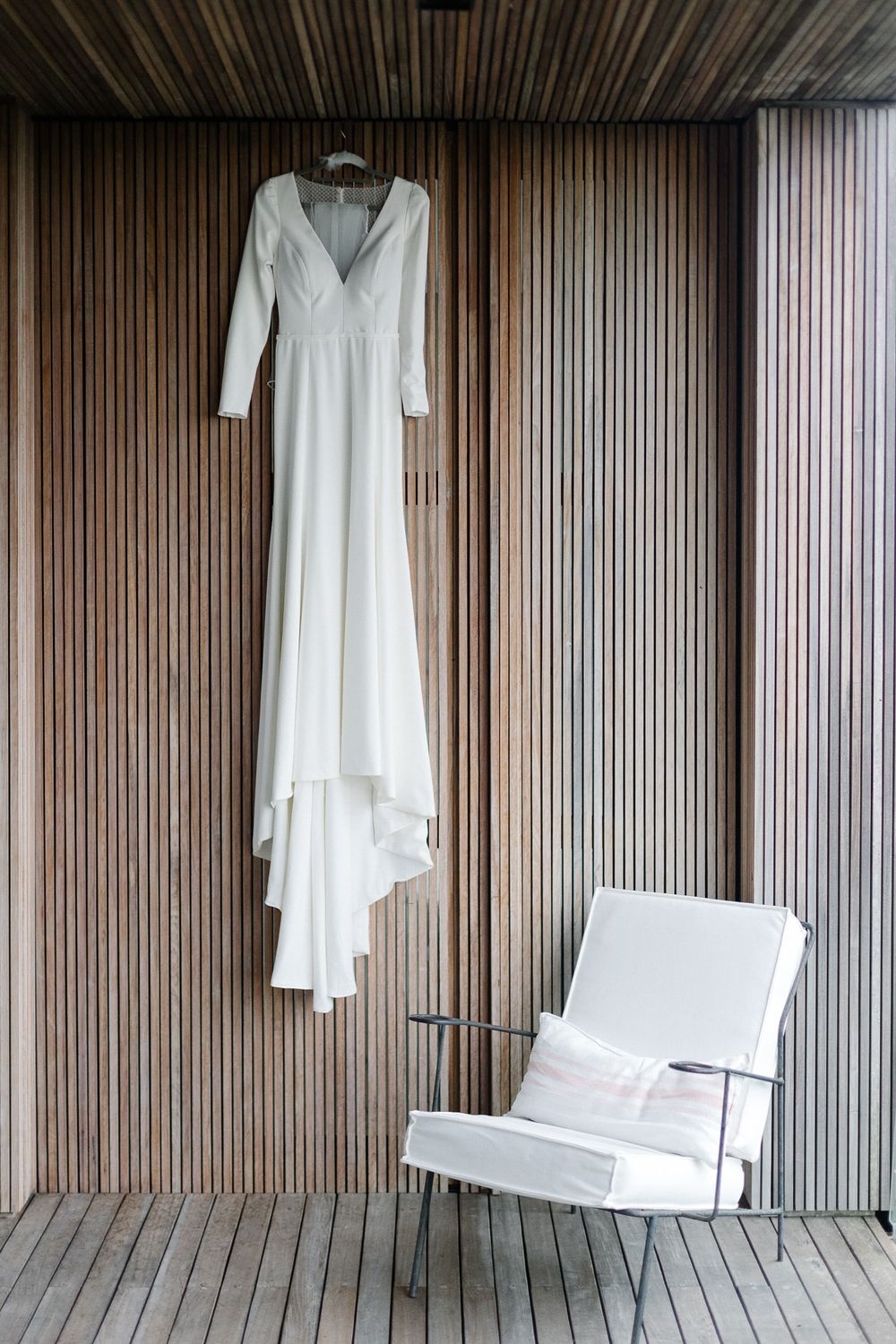 Elegant white wedding dress hanging on wooden paneled wall next to modern white chair.