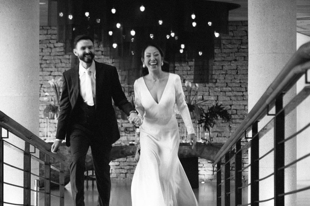 Joyful couple running through a brick-walled corridor lit with string lights.